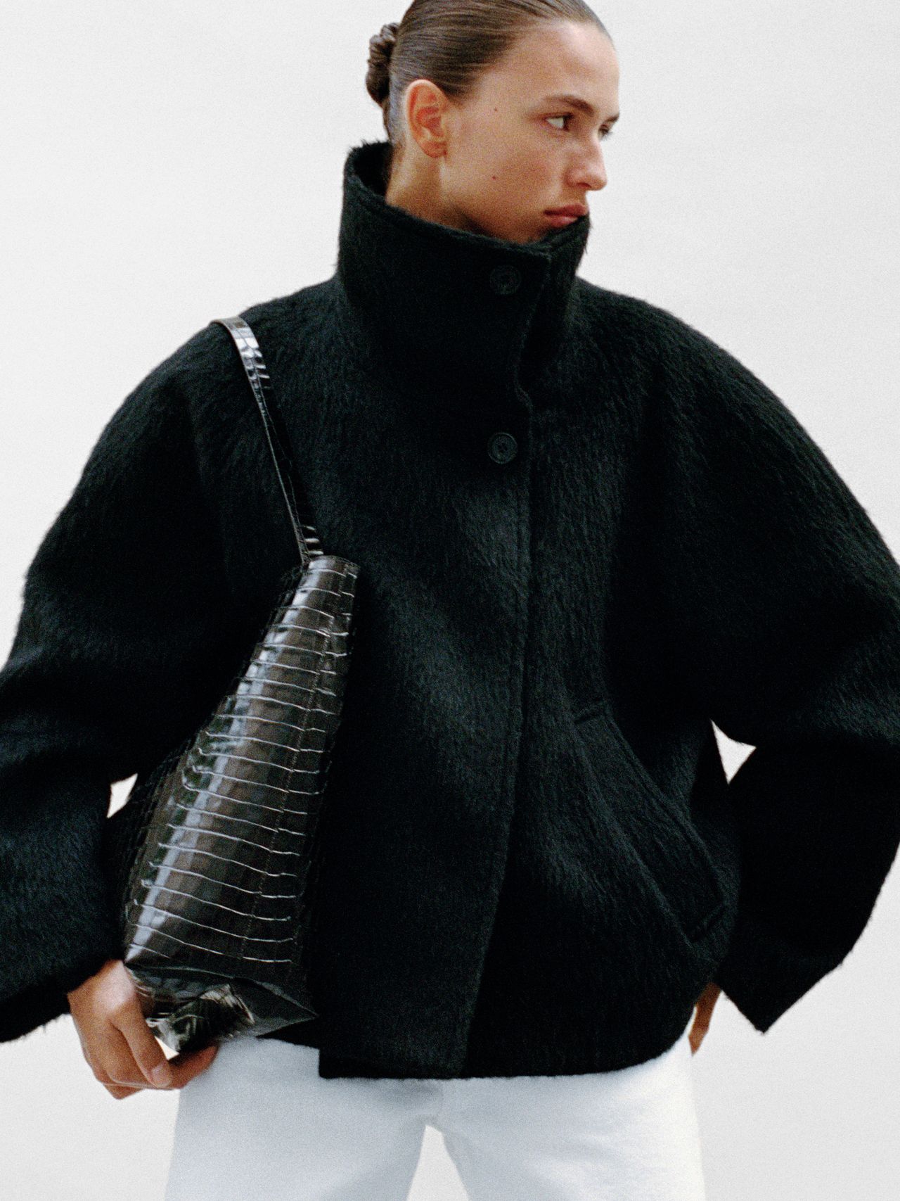 Person in a black textured coat and white pants, holding a large, black crocodile-patterned bag, with hair tied back, gazing to the side.