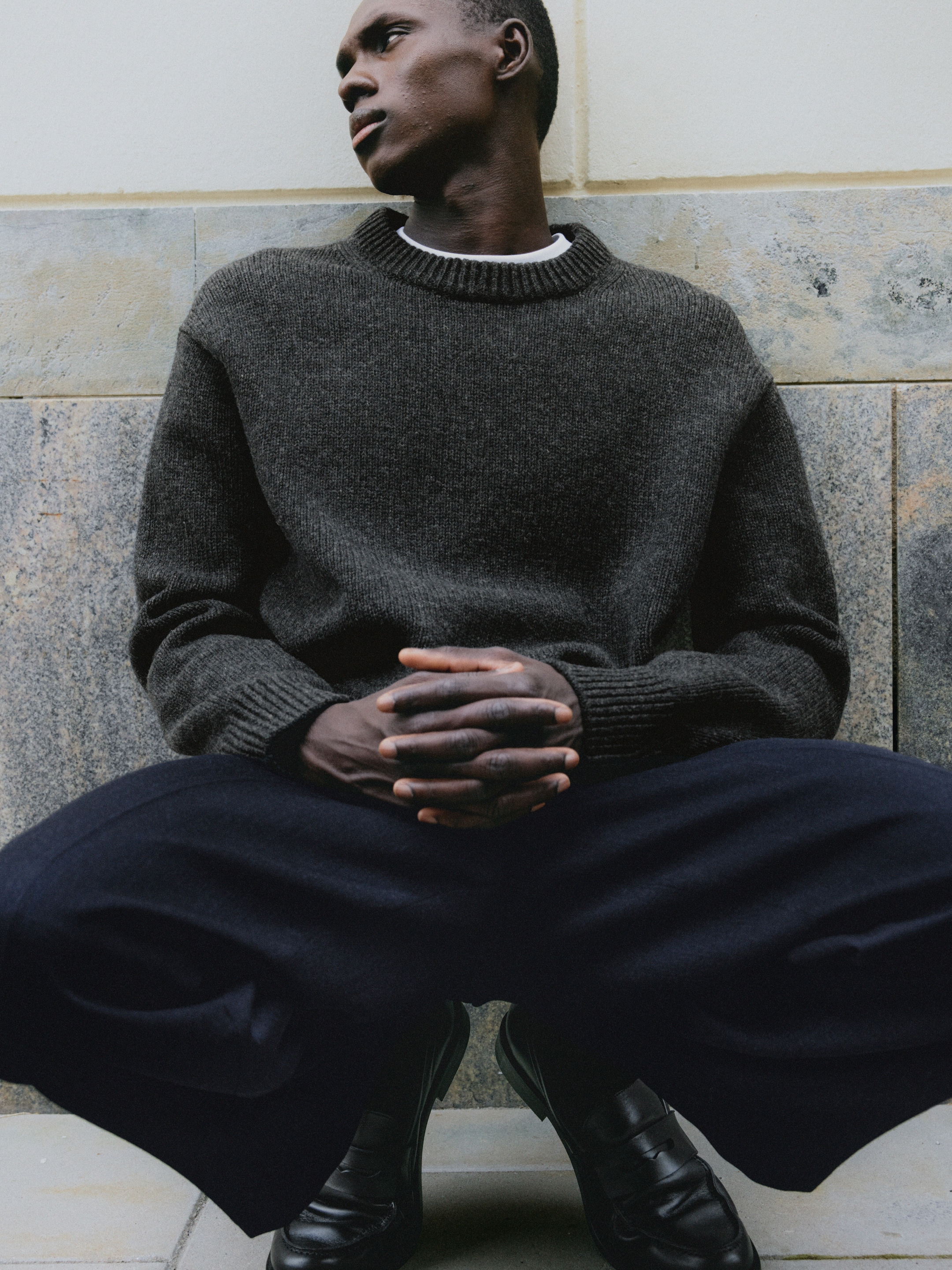 Person seated against a tiled wall, wearing a dark sweater and pants, looking to the side with hands clasped, exuding a contemplative mood.