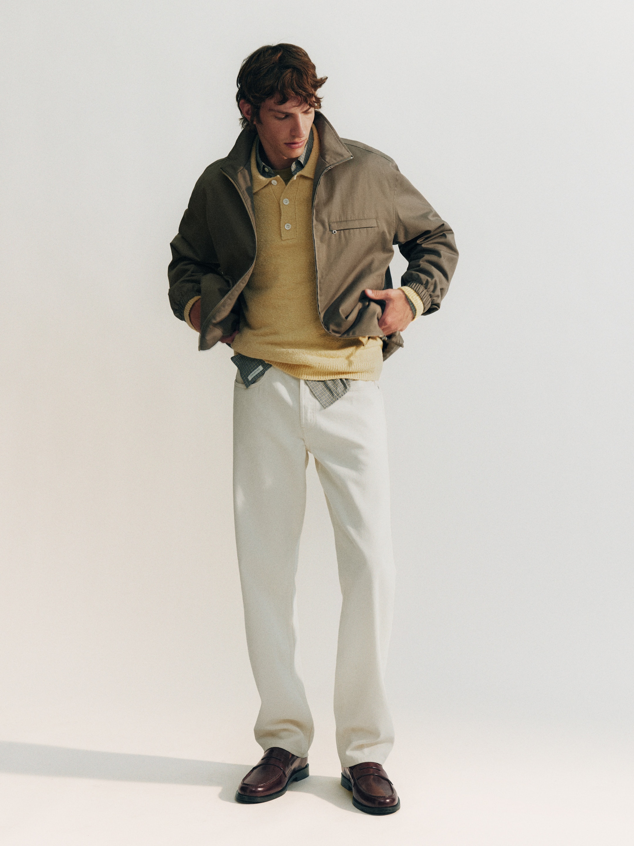 Man in beige jacket, yellow sweater, and white pants, adjusting his jacket. Brown loafers complete the casual look against a plain background.