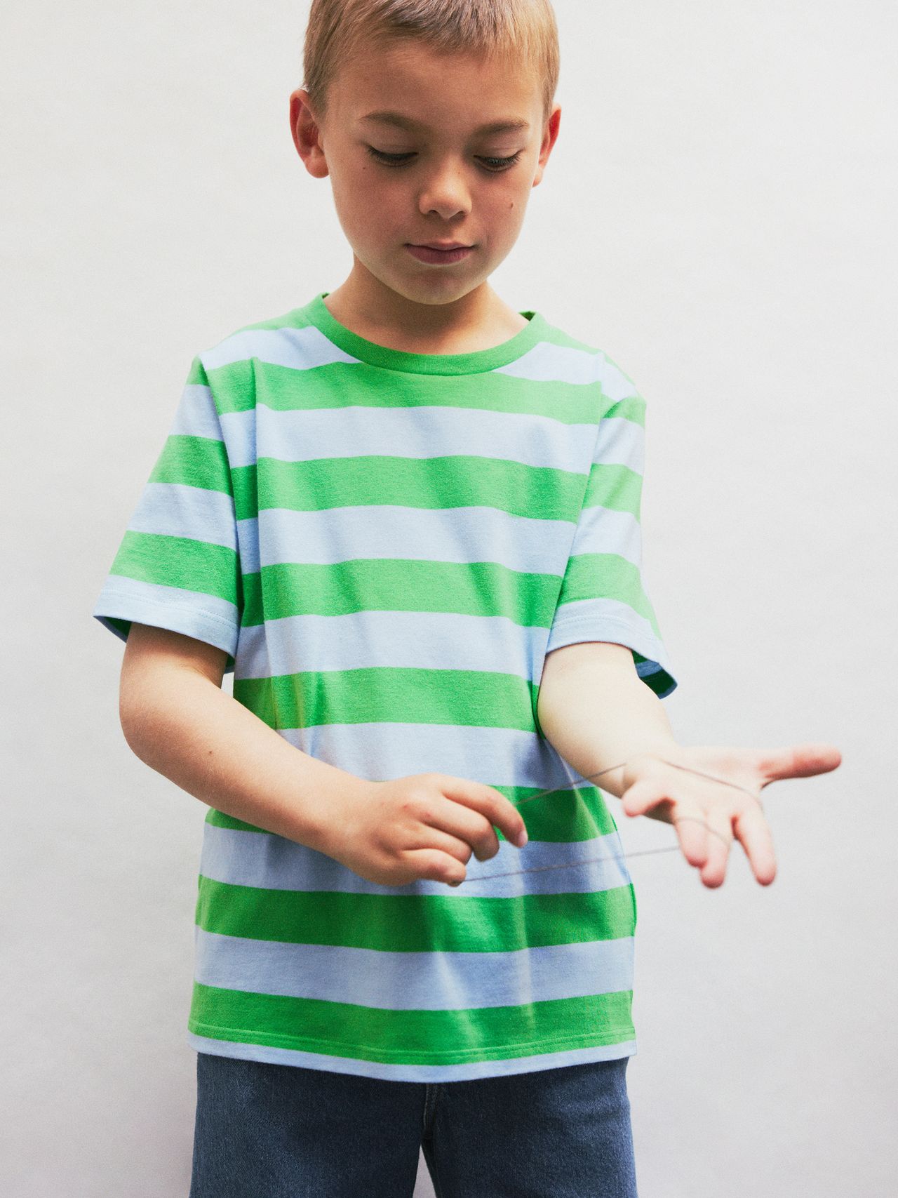 Young boy in a green and blue striped shirt playing with a rubber band, standing against a plain white background.