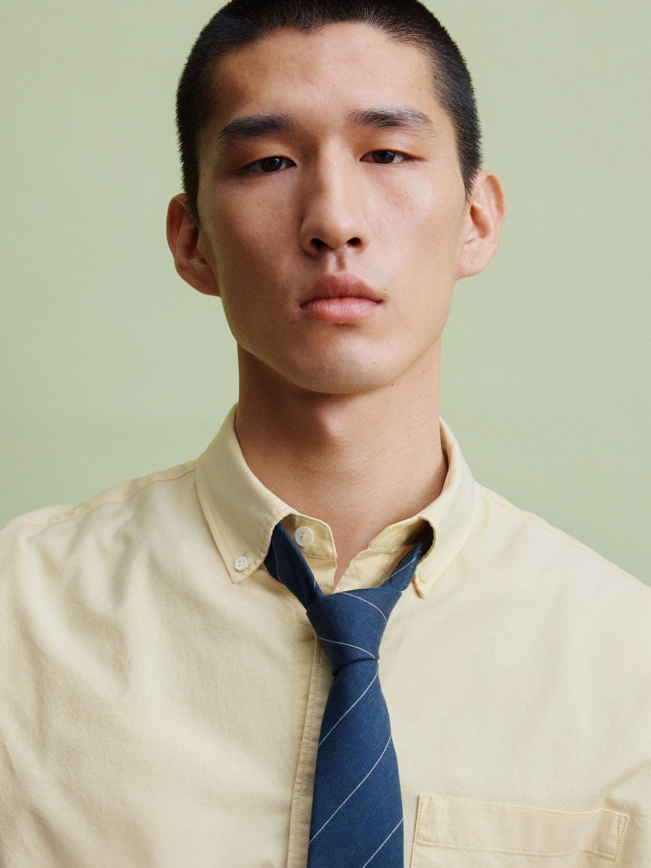 A person with short hair wearing a yellow button-up shirt and a loosely tied blue striped tie against a light green background.