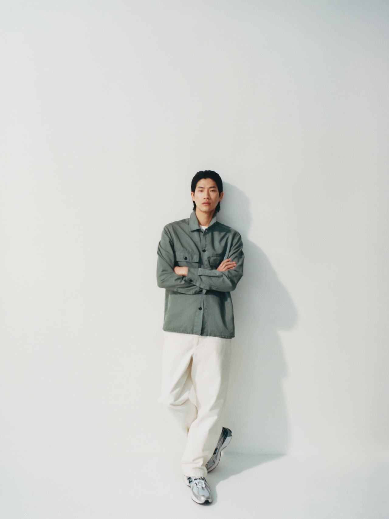 Person in a green button-up shirt and white pants stands against a white wall, arms crossed, with a relaxed expression.