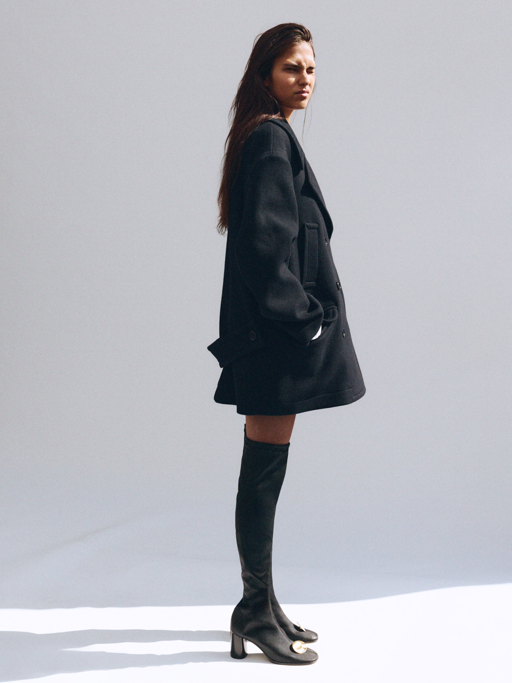 A person wearing an oversized black coat and knee-high black boots stands confidently against a plain backdrop.