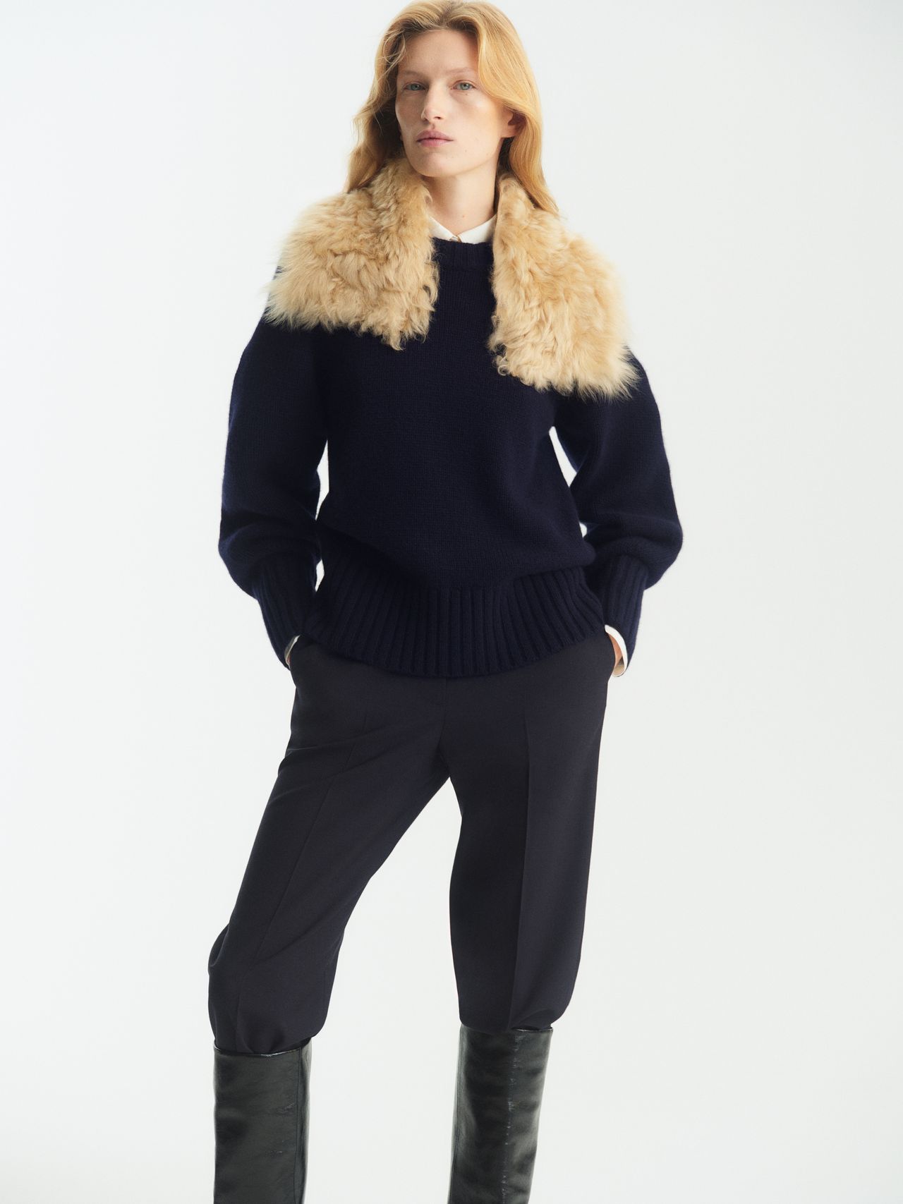 Person wearing a navy sweater with a fur collar, black pants, and black boots, standing against a plain white background.