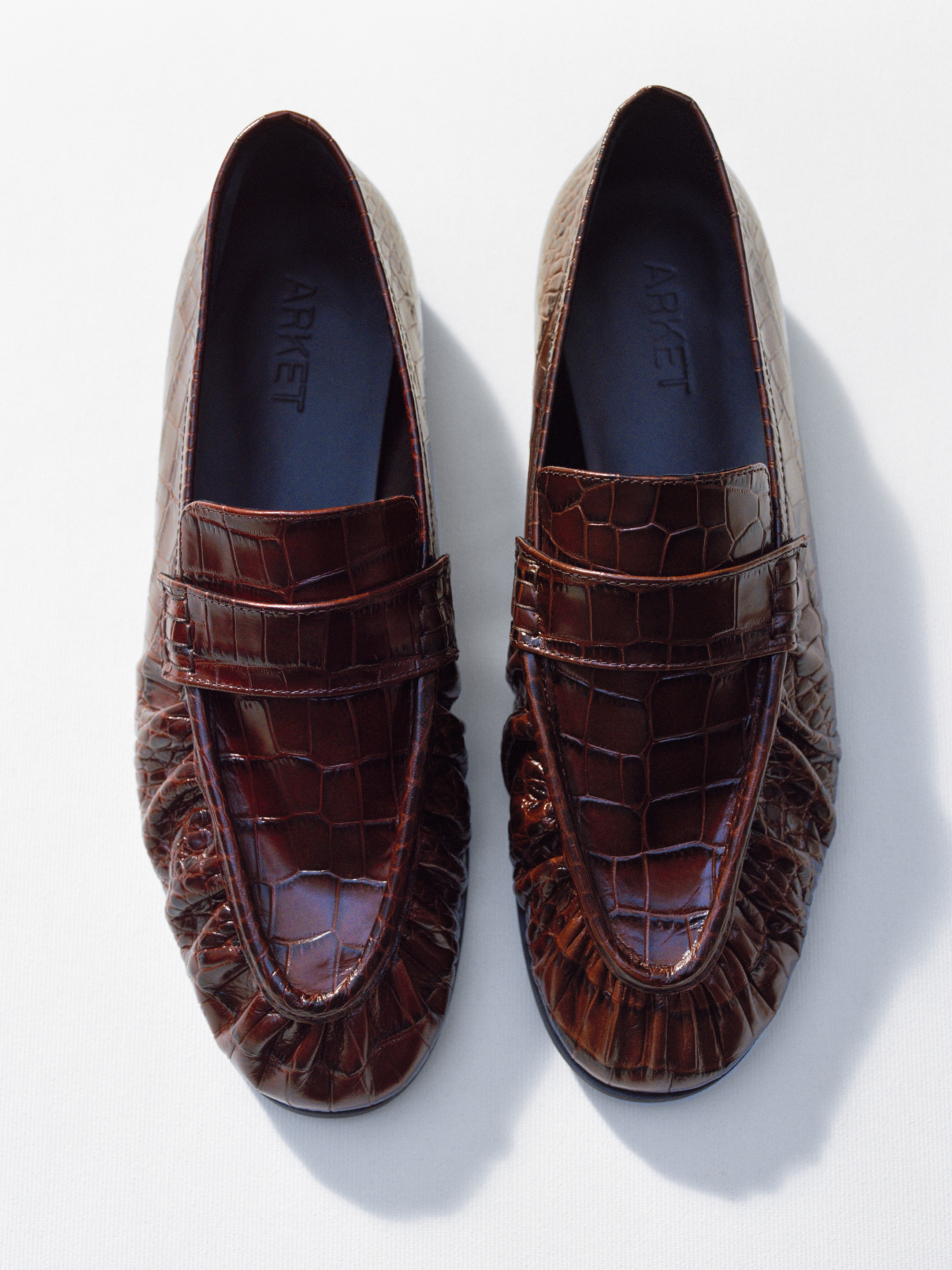 A pair of brown crocodile-textured leather loafers with pleated details, viewed from above against a white background.