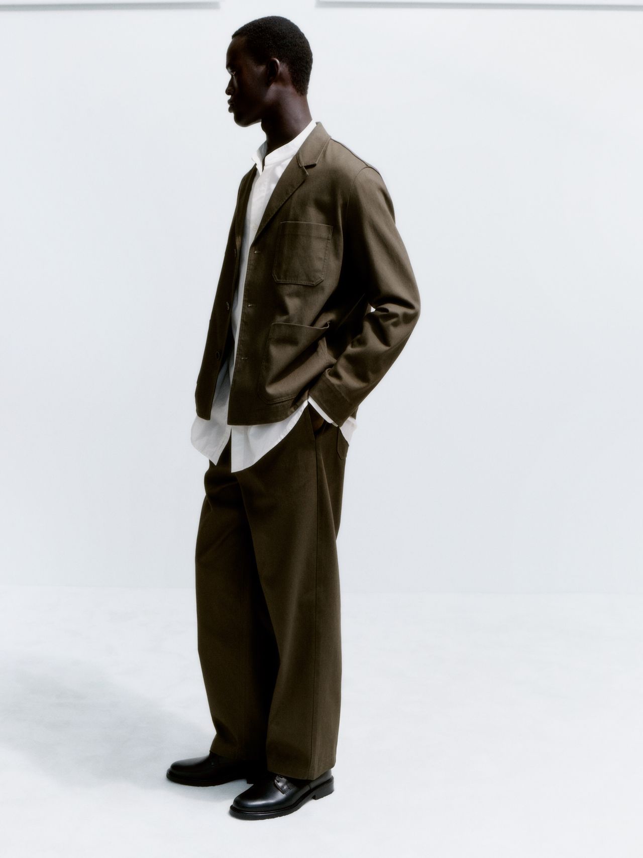 Man in a stylish khaki suit and white shirt, standing side profile against a plain white background, with hands in pockets.