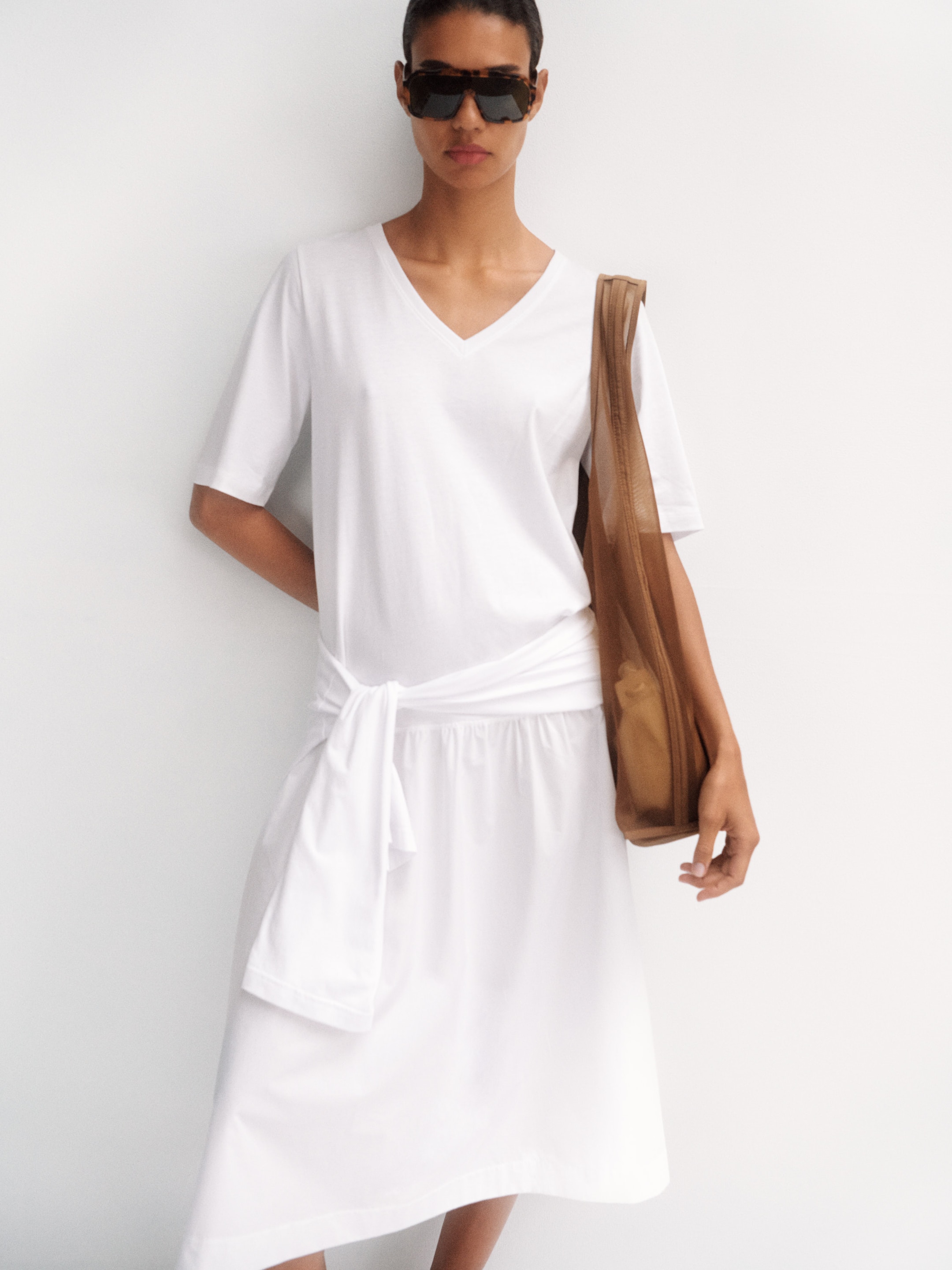 Person wearing a white outfit with sunglasses, holding a brown bag. The shirt has short sleeves and a V-neck. They pose against a white background.