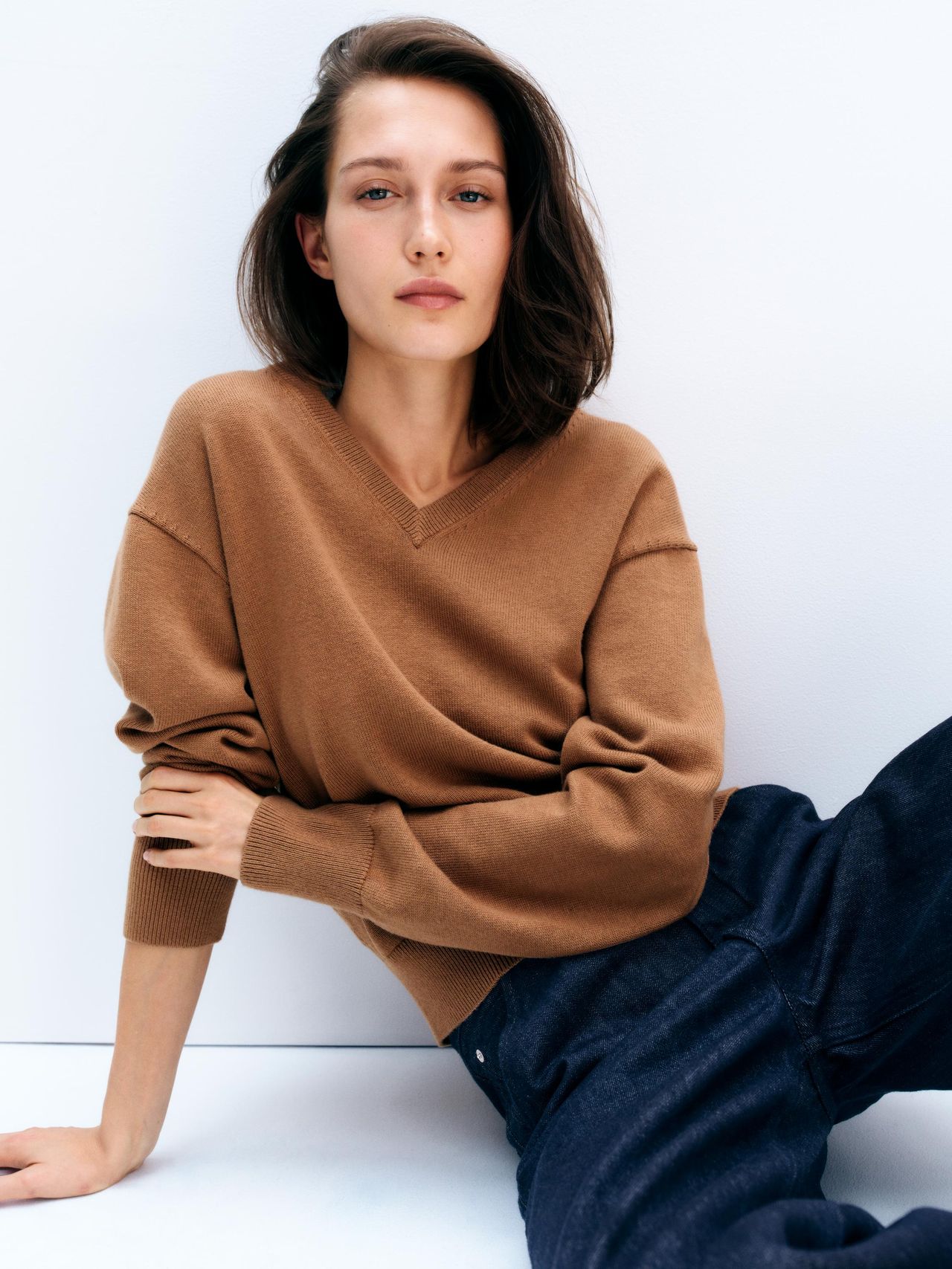Person sitting against a white wall, wearing a brown sweater and dark jeans, with a neutral expression and short, dark hair.