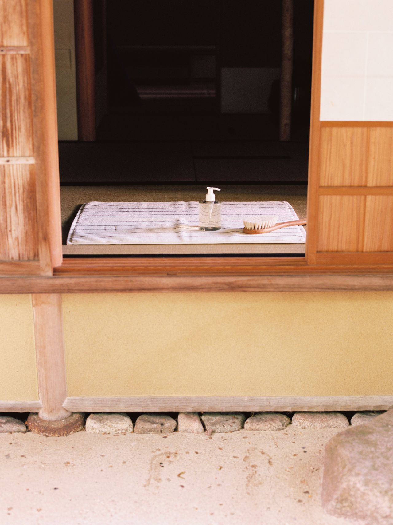 Open wooden door leading to a room with a mat, a bottle of soap, and a brush placed on it. Stone steps and sandy ground are visible outside.