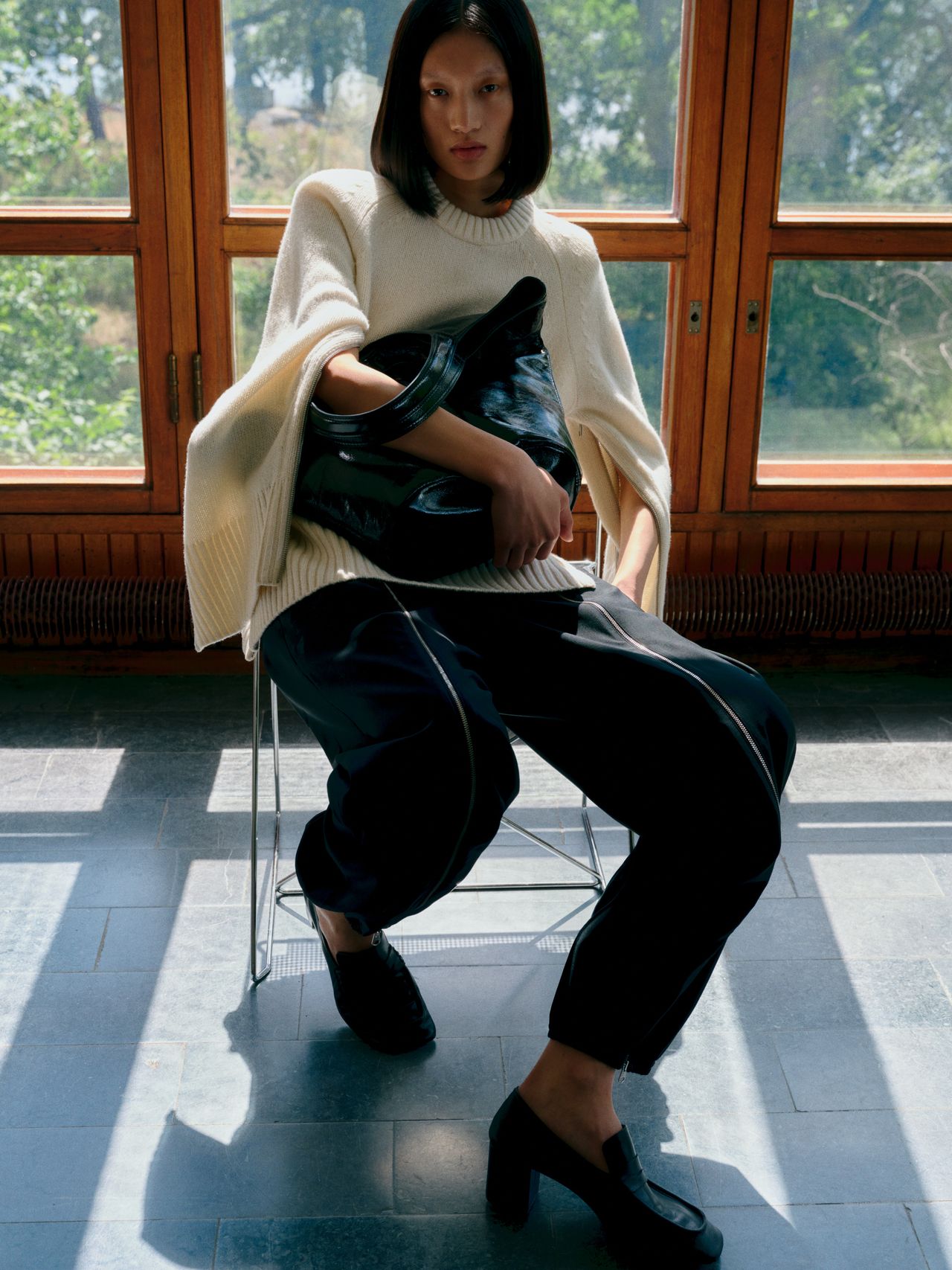 Woman in a white sweater and black pants sits on a chair, holding a black bag, with sunlight streaming through large windows behind her.
