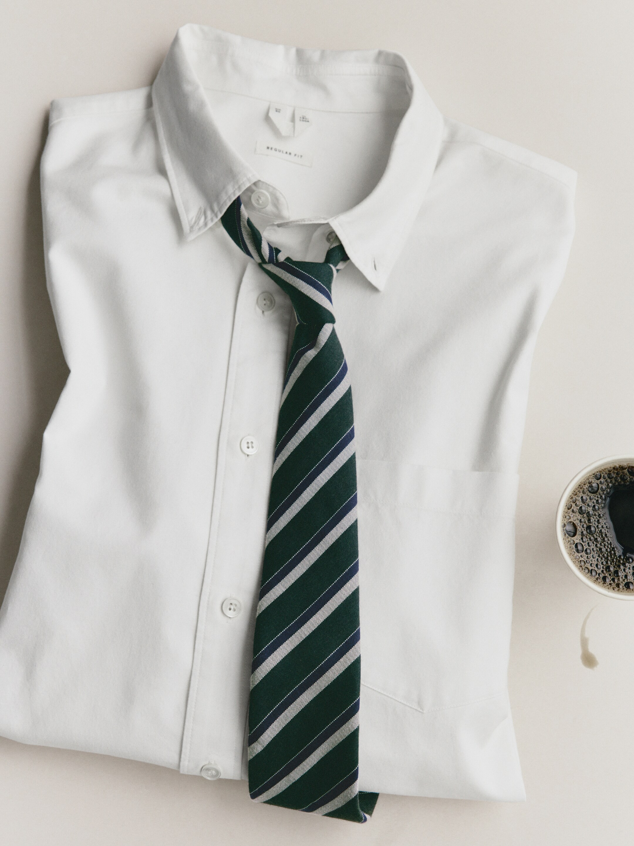 A folded white dress shirt with a dark green and navy striped tie, placed next to a cup of black coffee on a light surface.