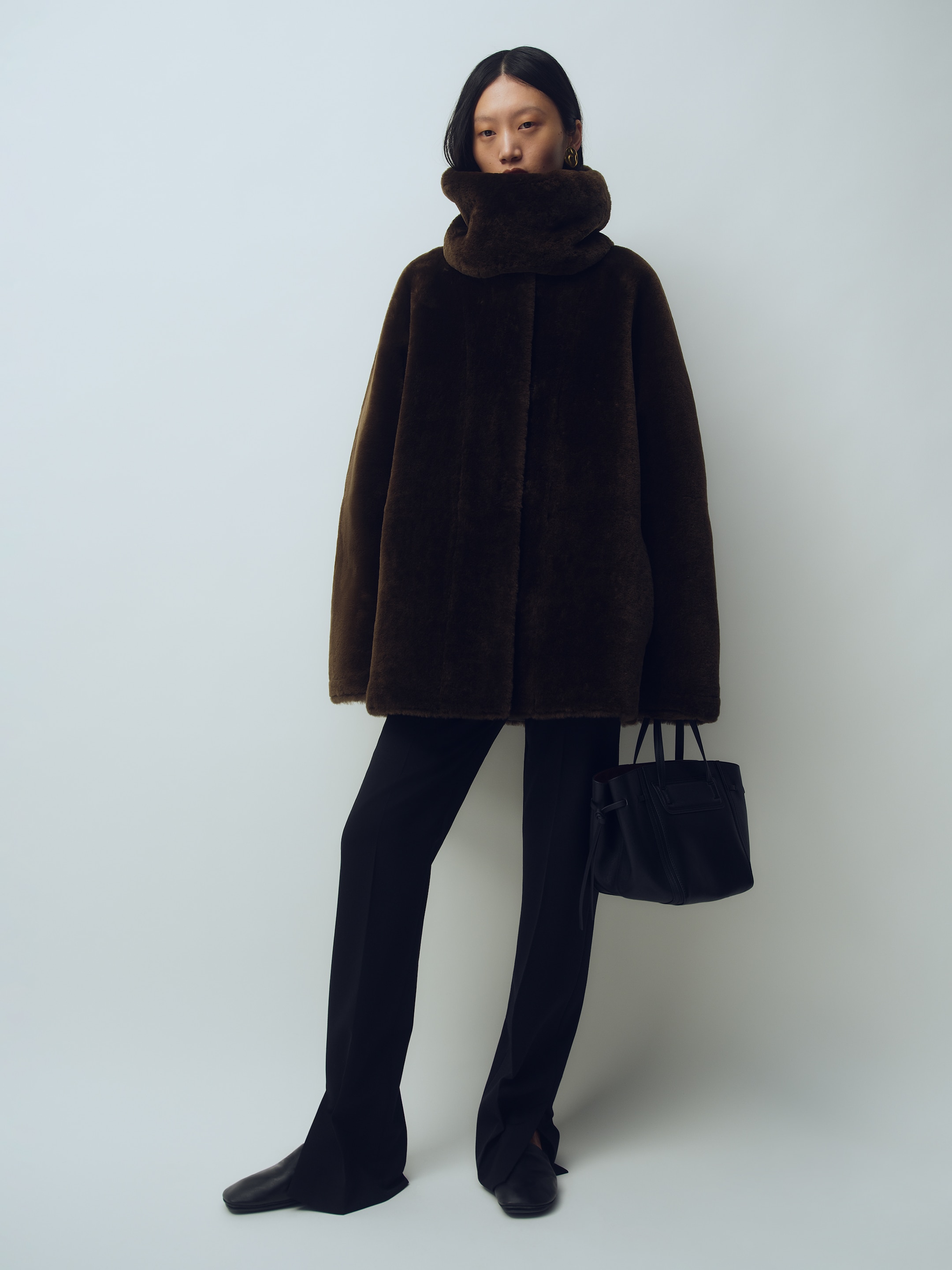 Person wearing a dark, oversized coat with a high collar, black pants, and holding a black handbag, standing against a plain background.