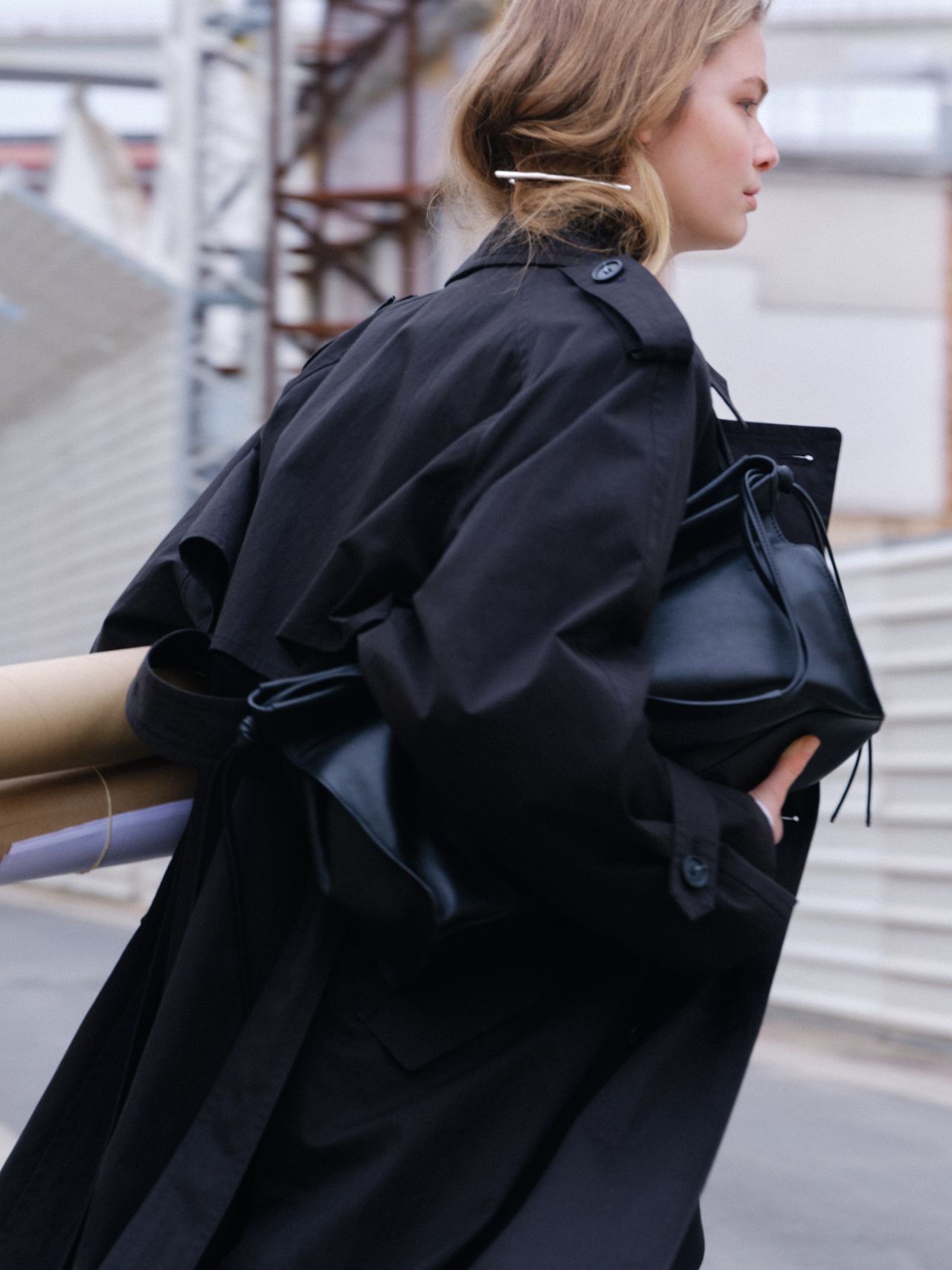 A person in a black trench coat carries rolled papers and a black bag, walking by a construction site with metal fencing.