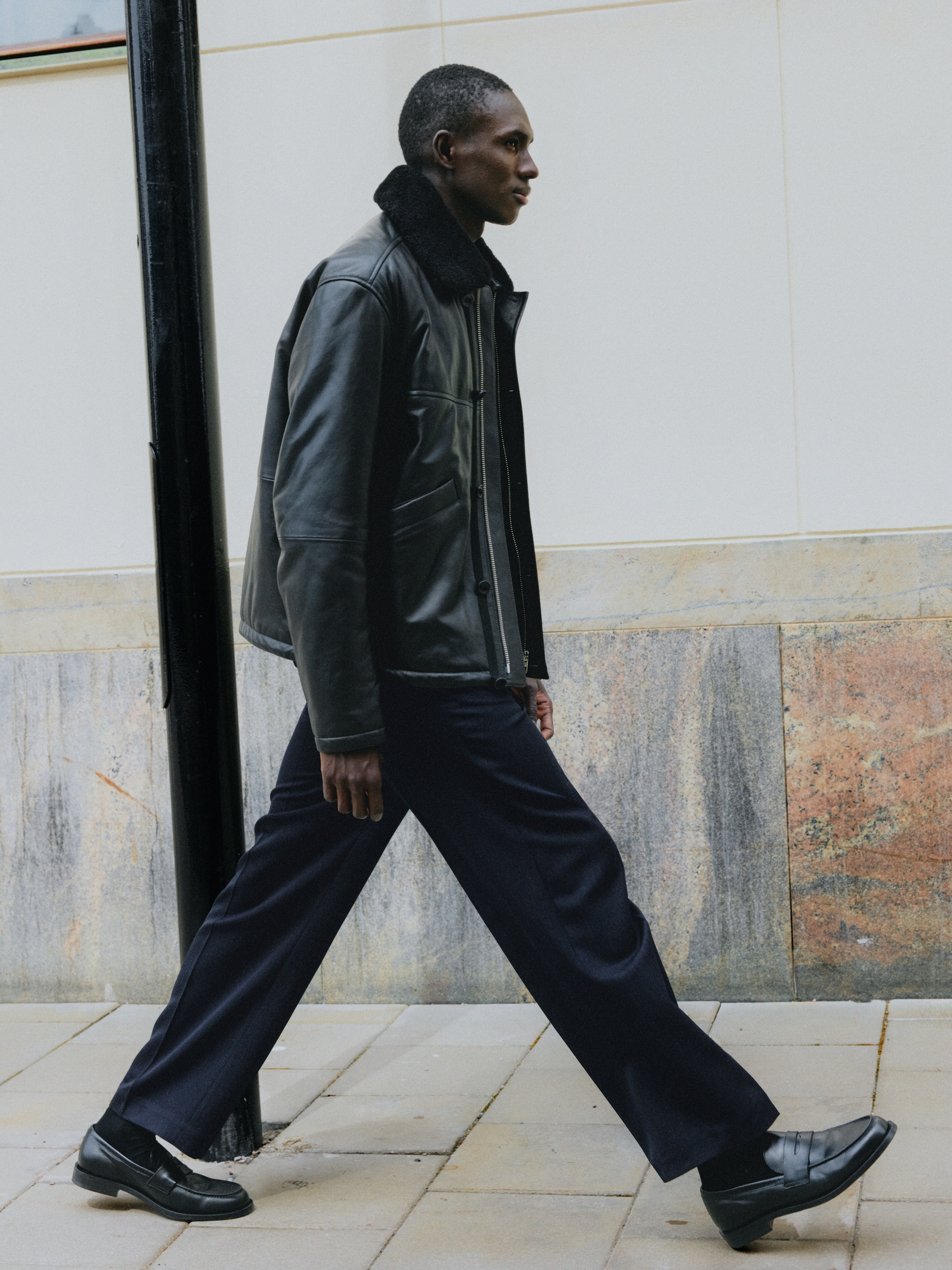 Man walking on pavement, wearing a black leather jacket, dark trousers, and black shoes, against a light wall background.