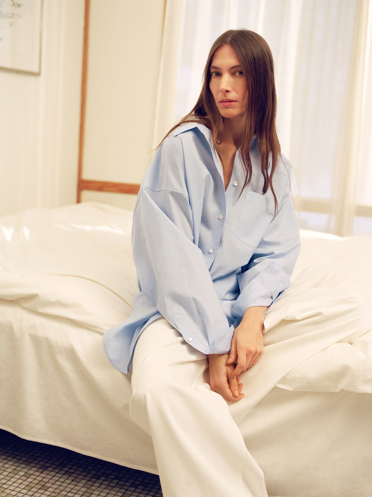 A woman with long hair sits on a white bed, wearing an oversized light blue shirt and white pants in a softly lit room.