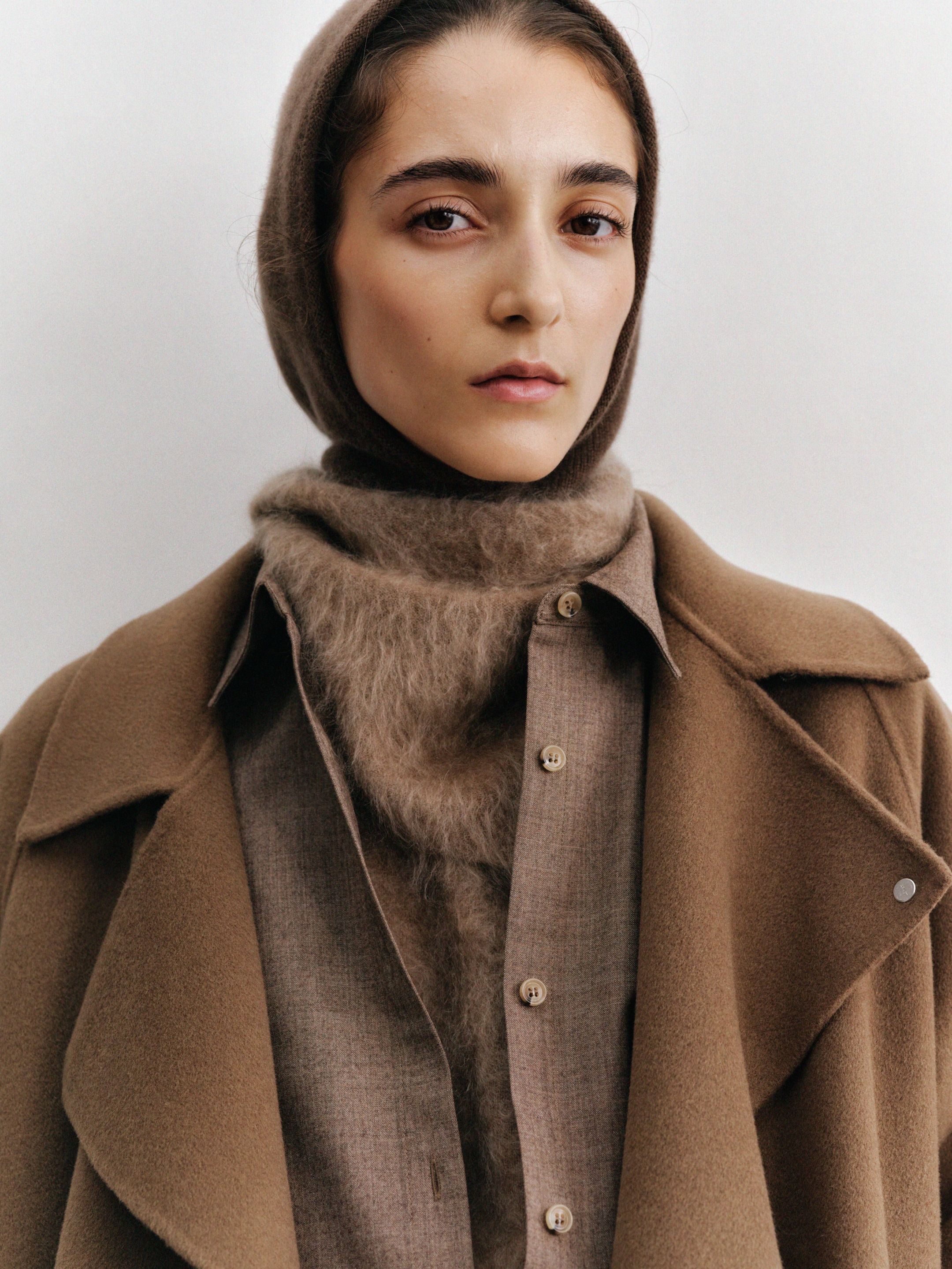 A person wearing a brown coat, matching scarf, and hood, looking directly at the camera against a neutral background.
