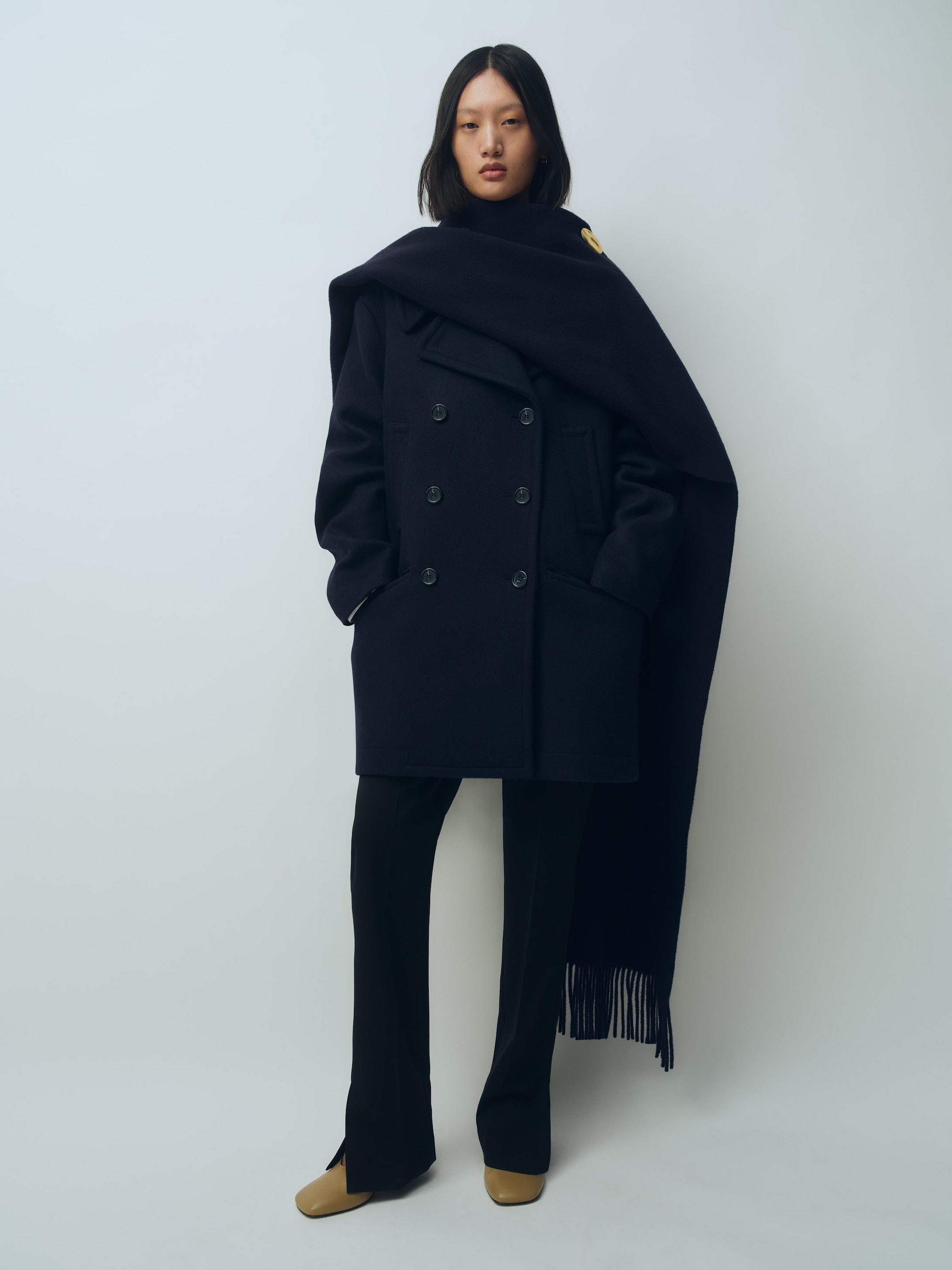A person wearing a dark double-breasted coat, matching scarf, black pants, and tan shoes stands against a plain white background.