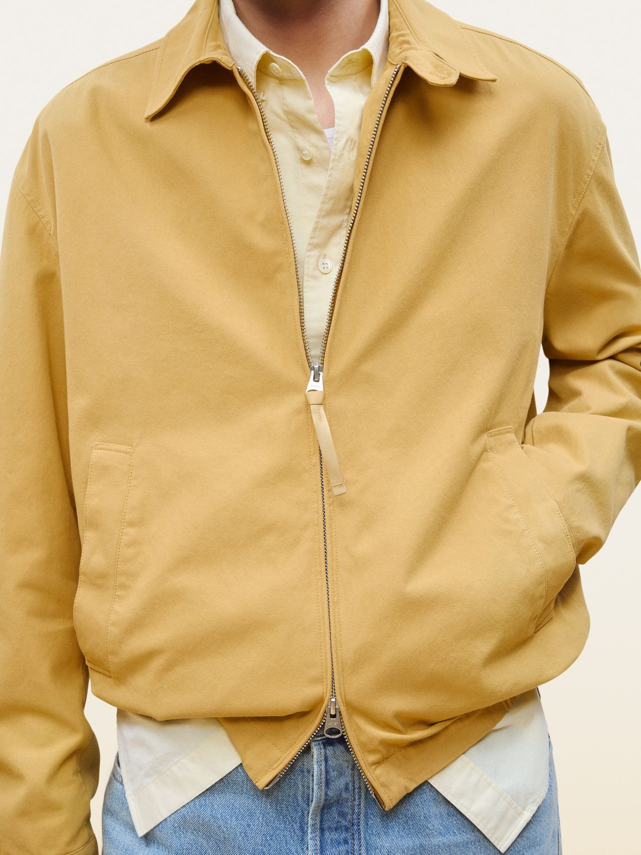 Person wearing a beige collared jacket over a light yellow shirt and blue jeans, with hands tucked in jacket pockets.