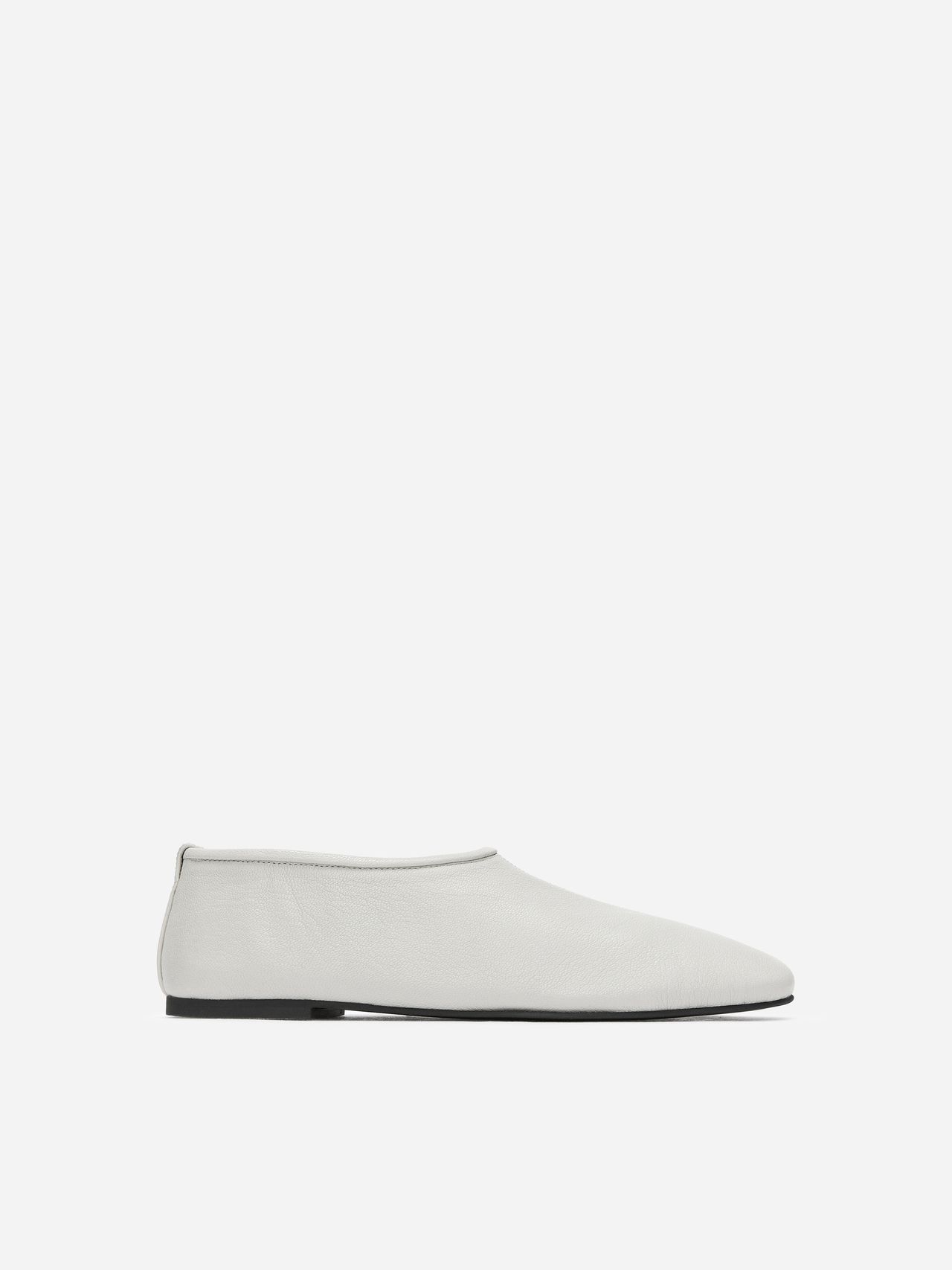 Minimalist white leather slip-on shoe with a flat black sole, displayed on a white background.