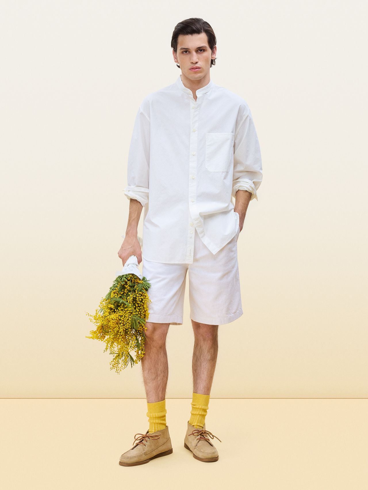 Man in a white shirt and shorts holds yellow flowers, wearing yellow socks and brown shoes, against a beige background.