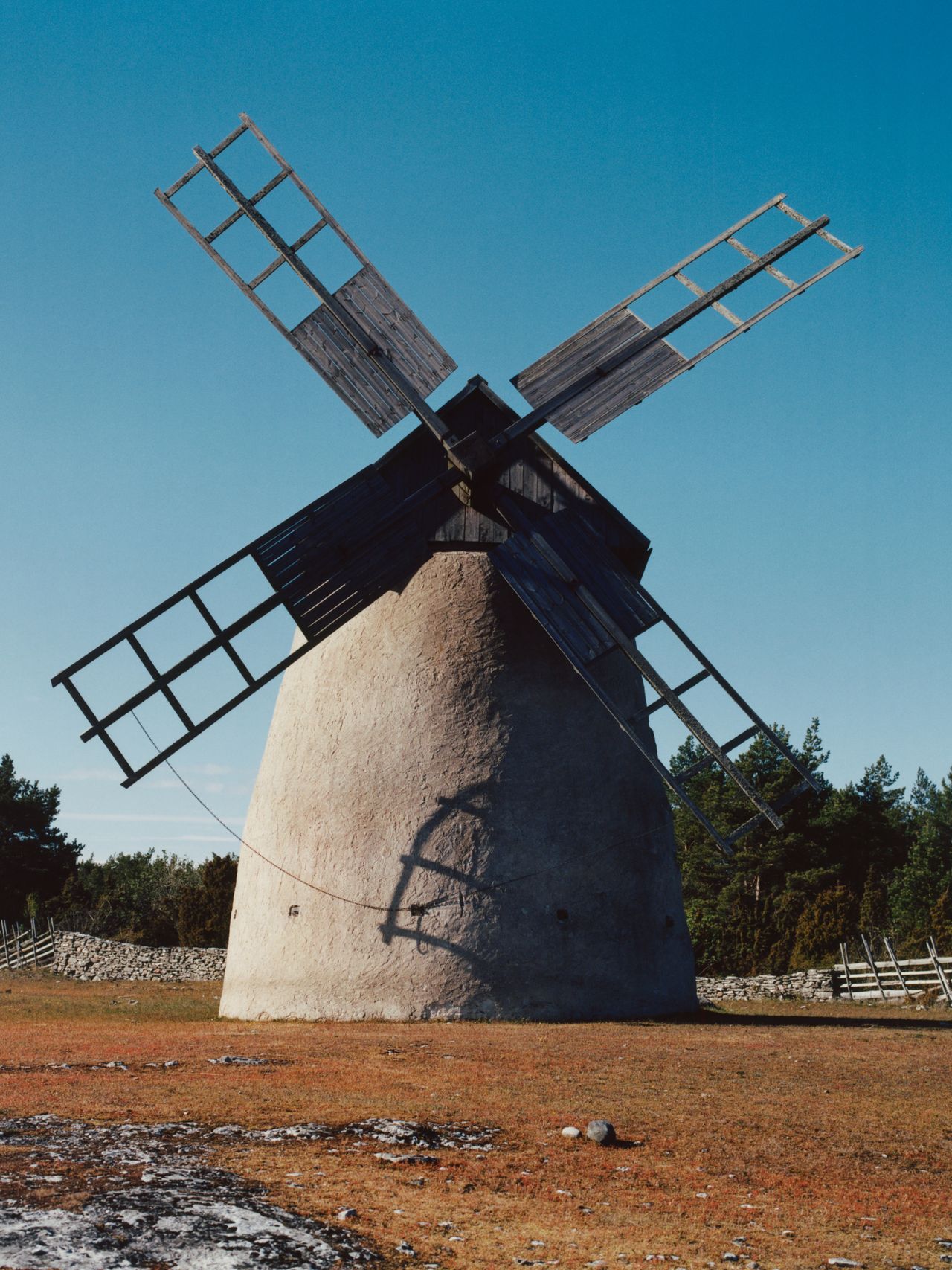 A rustic windmill with wooden blades stands on a grassy field under a clear blue sky, surrounded by trees and a stone fence.