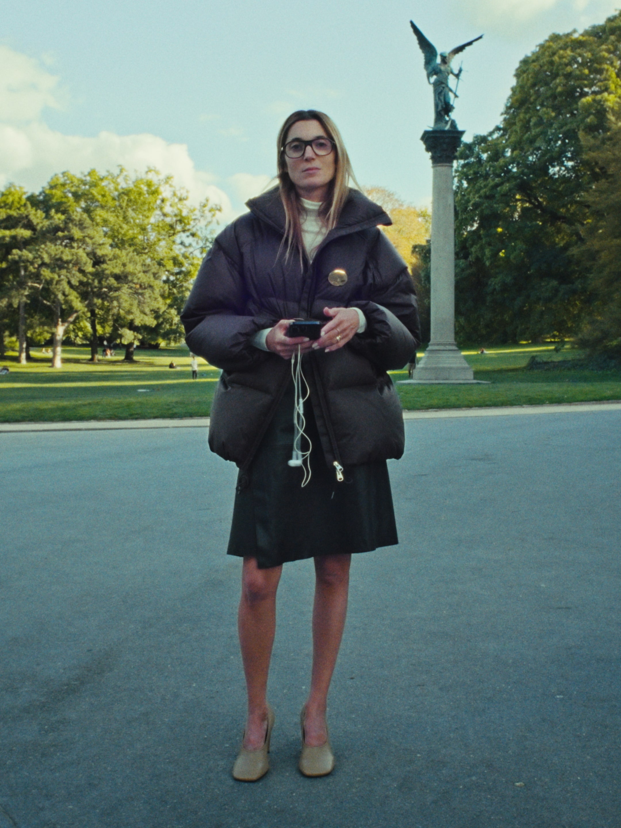 A woman stands in a park, wearing a dark puffer jacket and glasses, holding a phone. A statue on a column is visible in the background.
