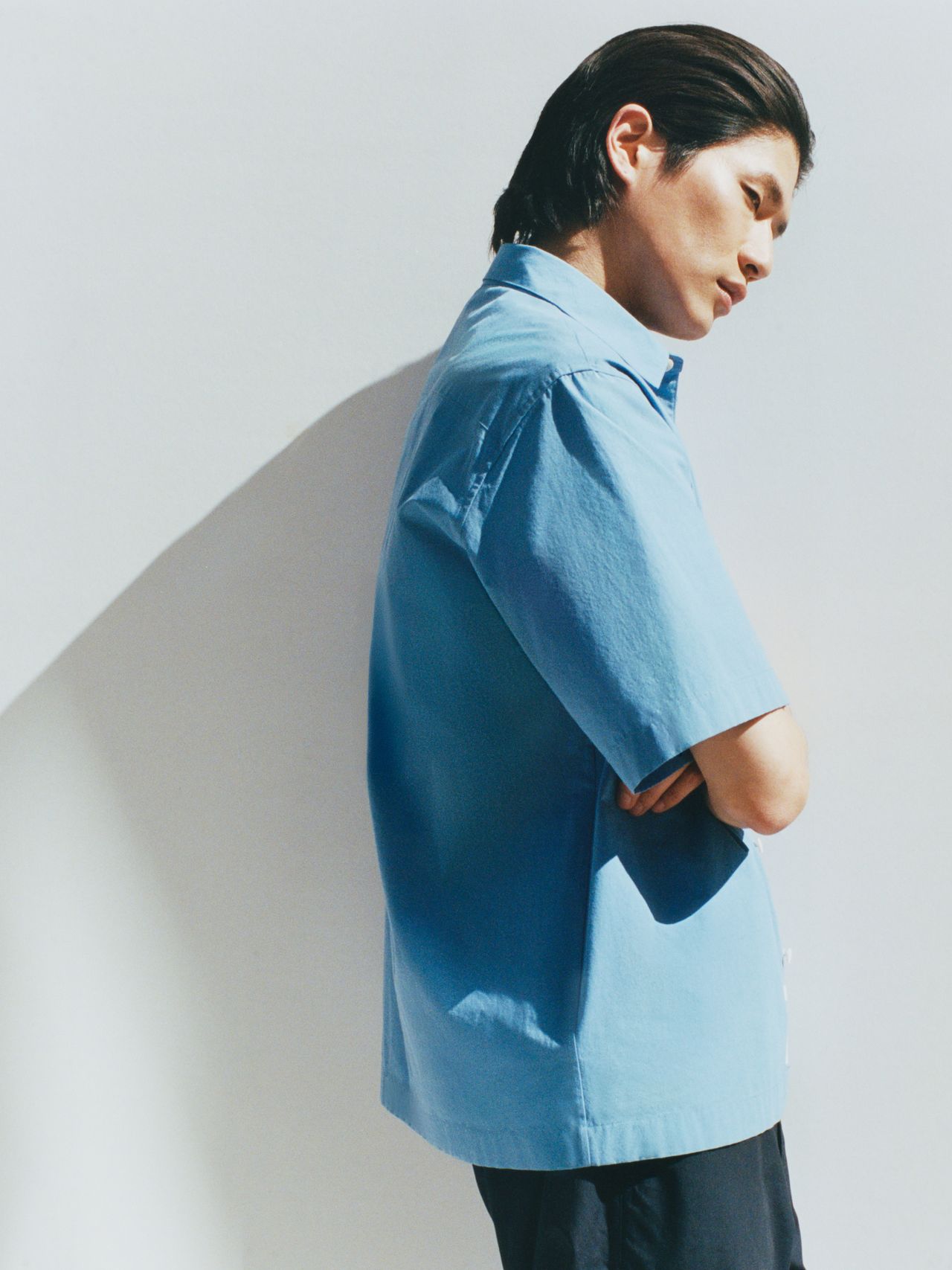 Person in a blue short-sleeve shirt stands against a white background, arms crossed, looking down. Shadows cast on the wall.