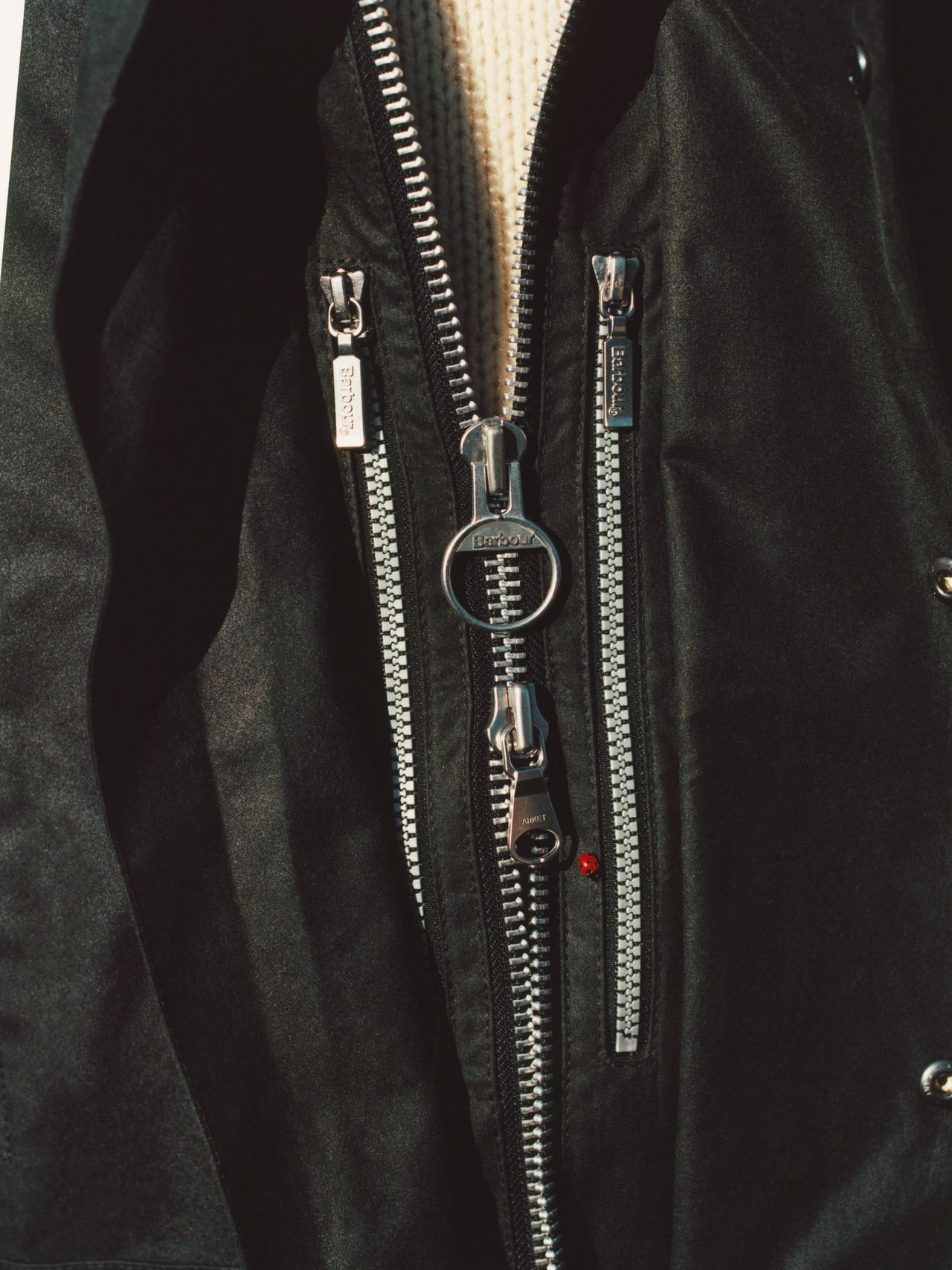 Close-up of a black jacket with multiple silver zippers, including a large central zipper with a circular pull, revealing a white fabric underneath.