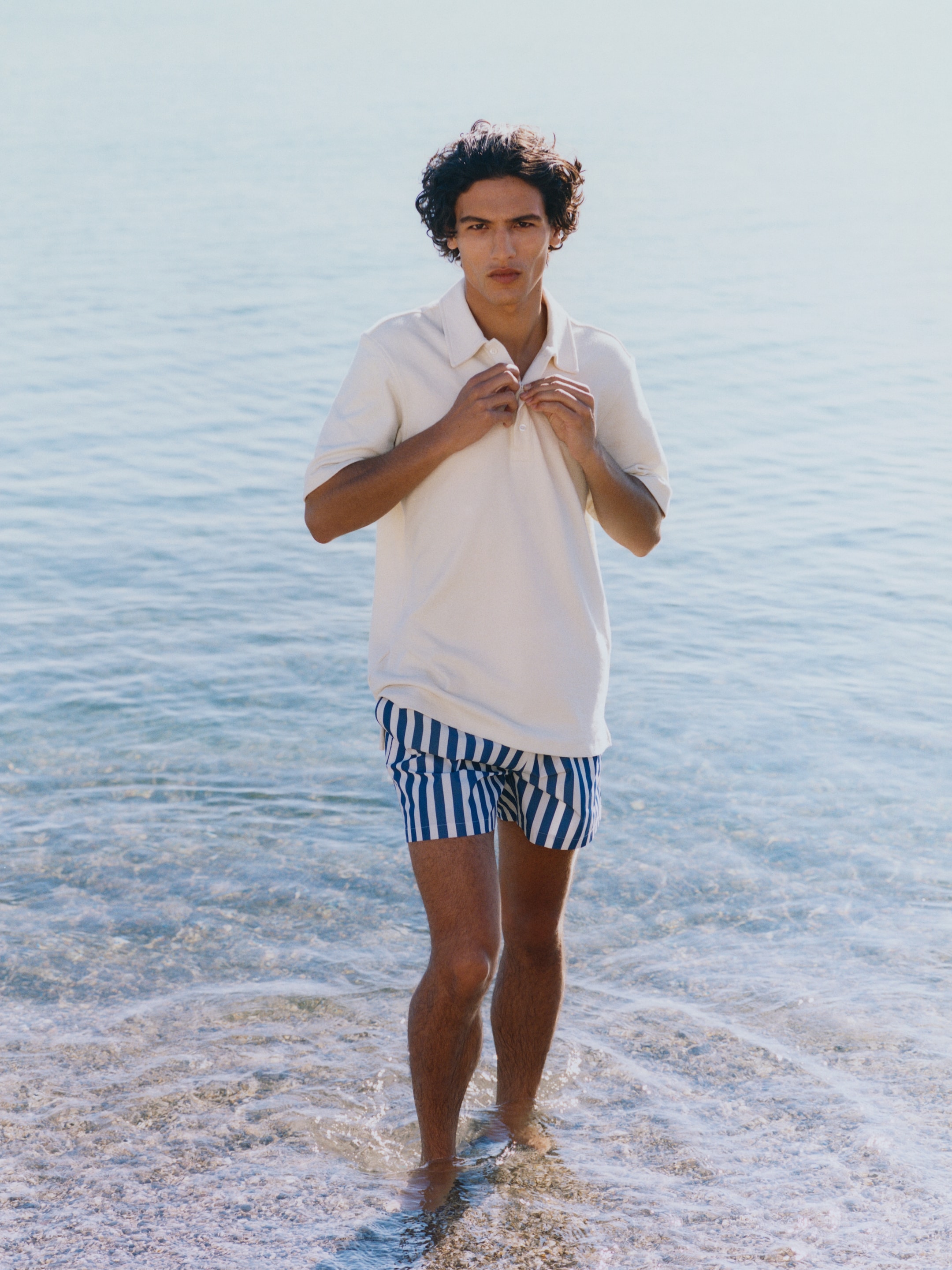Person standing in shallow water at the beach, wearing a white shirt and blue-striped shorts, with a calm sea in the background.