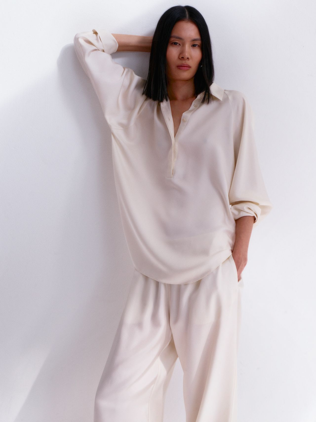 Person in a loose, cream-colored outfit poses against a white wall, with one arm resting on the wall and a hand in their pocket.