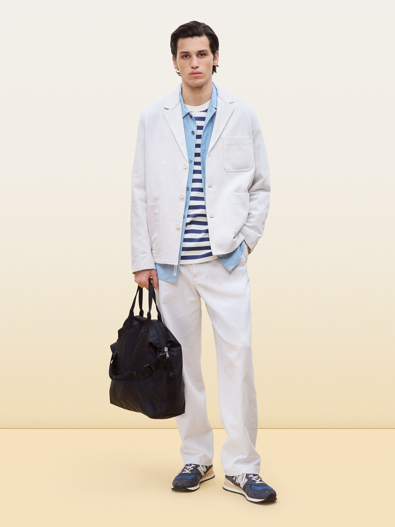 Man in a light-colored suit with a striped shirt, holding a black bag, and wearing blue sneakers, standing against a neutral background.