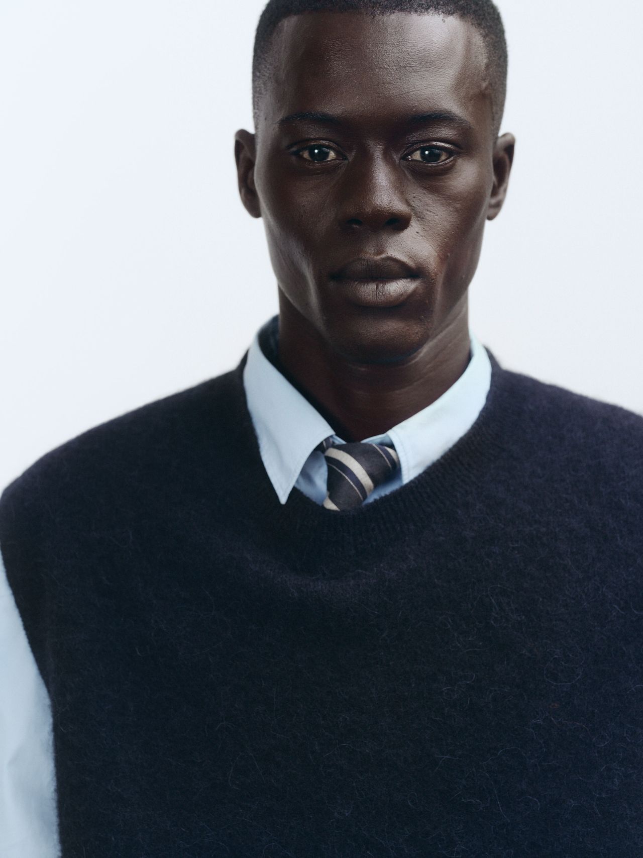 A person wearing a navy sweater over a light blue shirt and striped tie, standing against a plain white background.