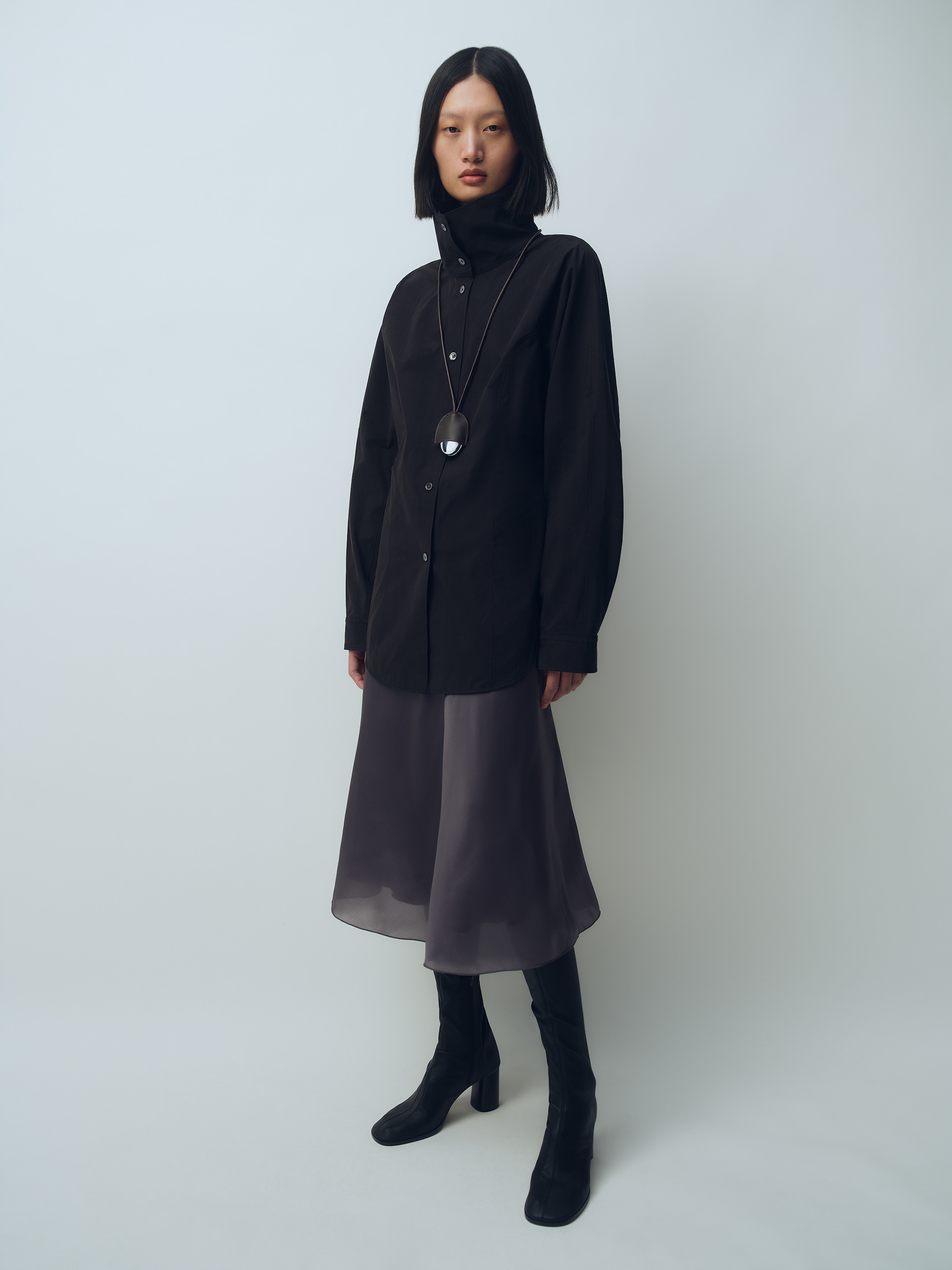 A person with shoulder-length hair wears a long black shirt, gray skirt, and tall black boots, accessorized with a long pendant necklace.