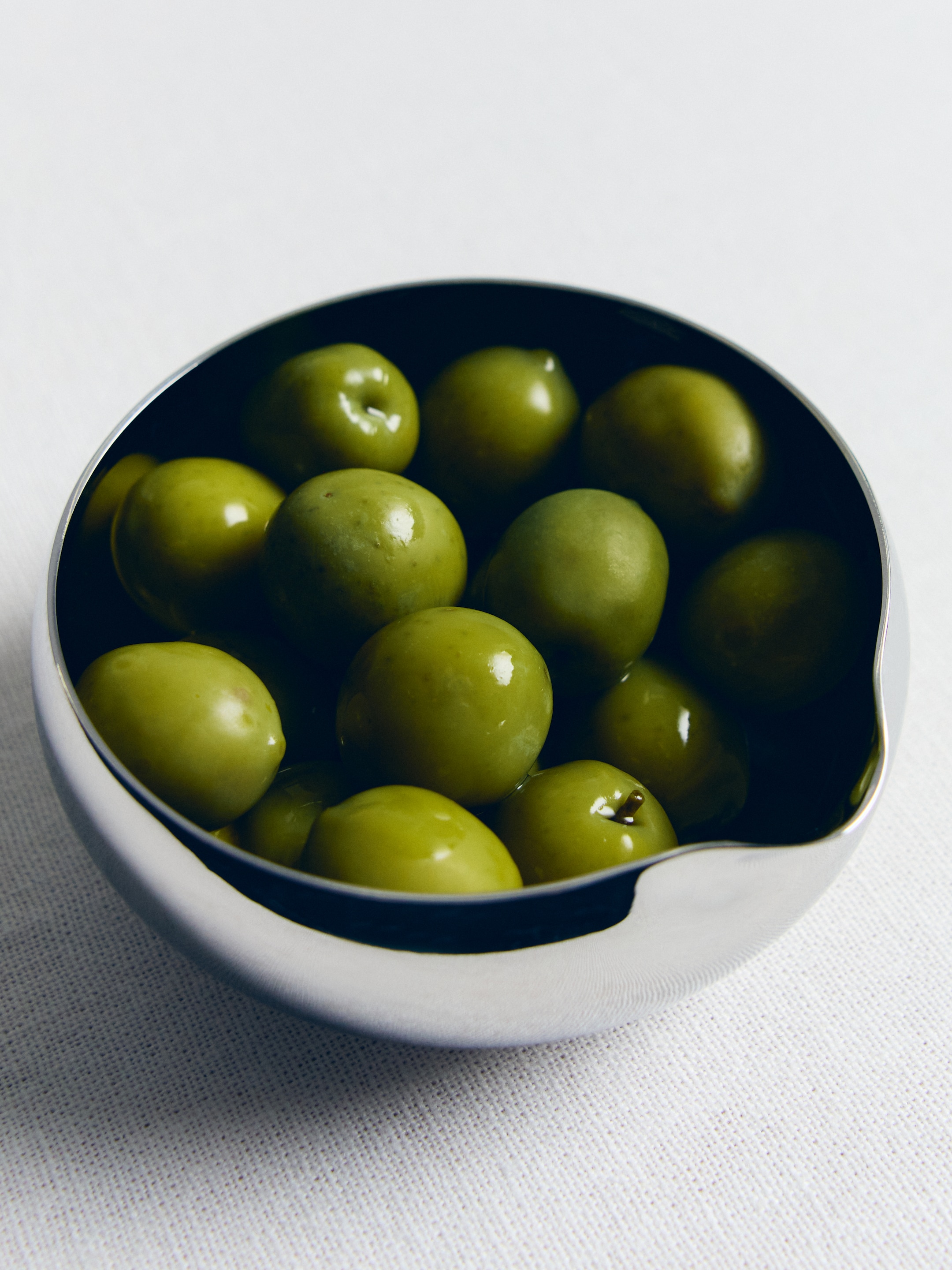 A shiny silver bowl filled with glossy green olives on a white surface.