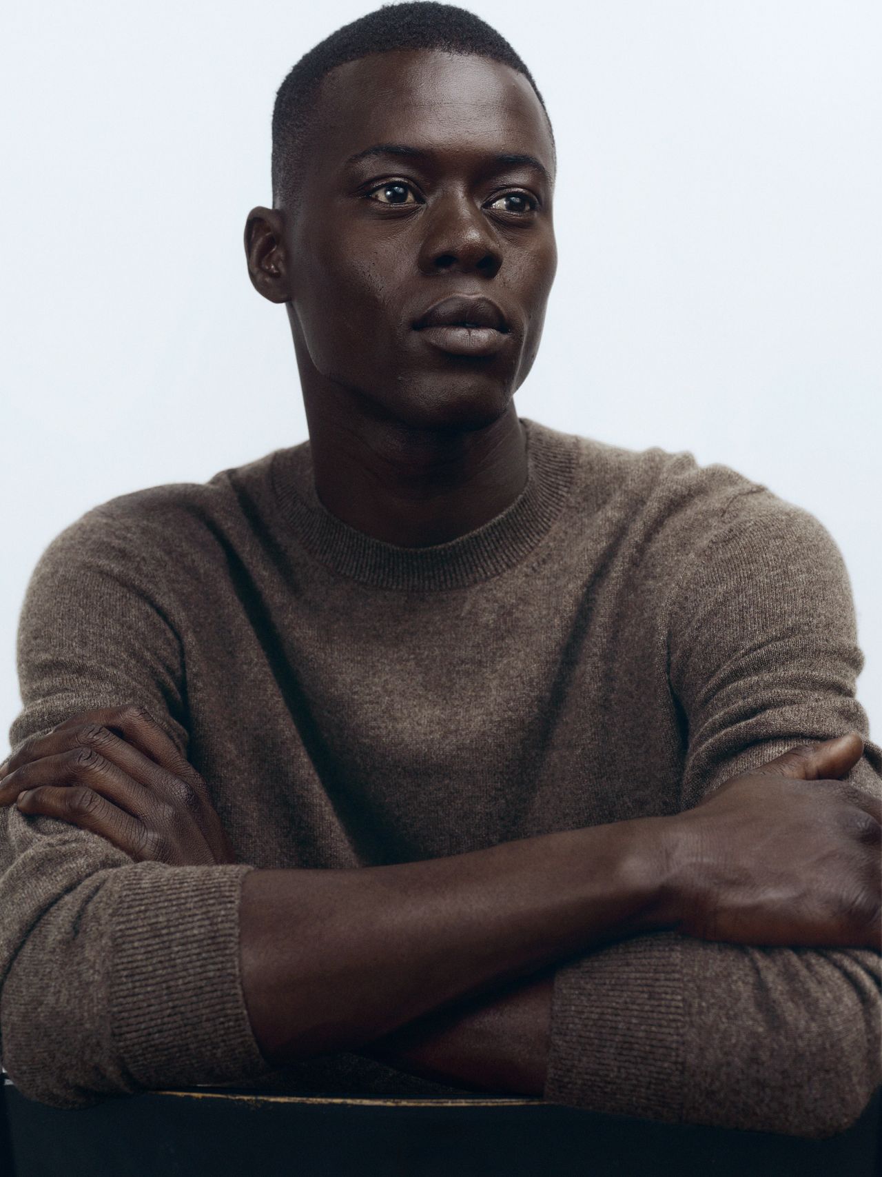 Person with short hair in a brown sweater, sitting with arms crossed, looking slightly to the side against a plain background.