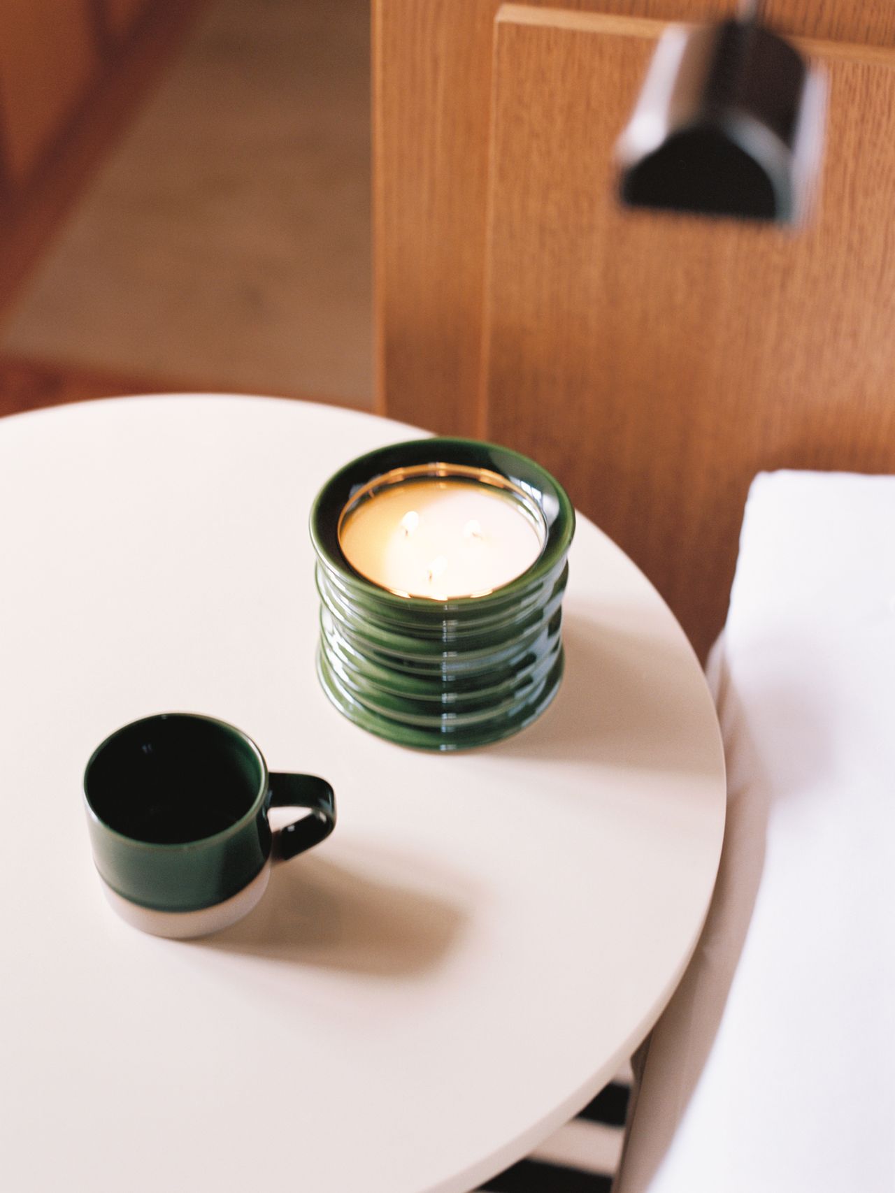 A lit candle and a dark green cup sit on a small, round white table near a wooden wall.