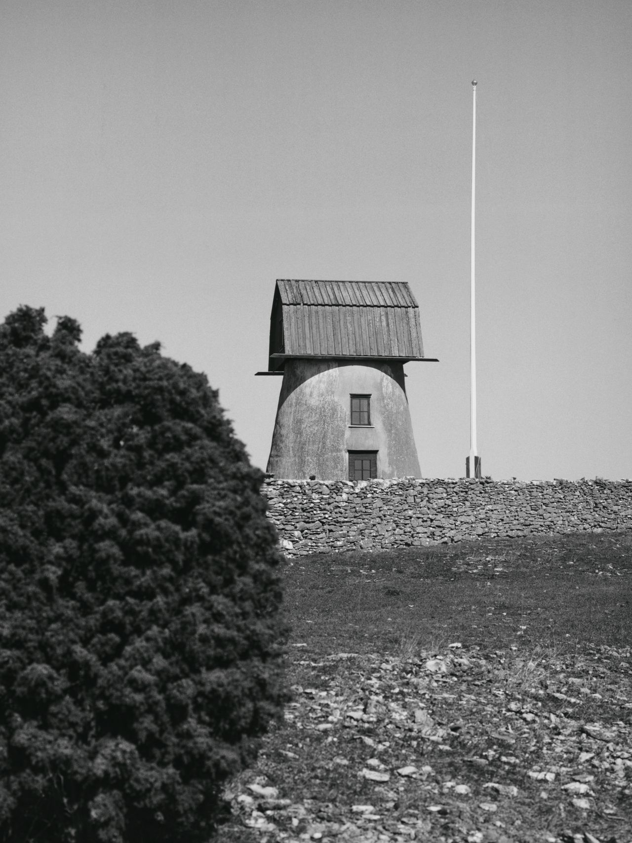 Black and white image of a windmill with a wooden roof, stone base, and nearby tall flagpole, framed by a bush and rocky ground.