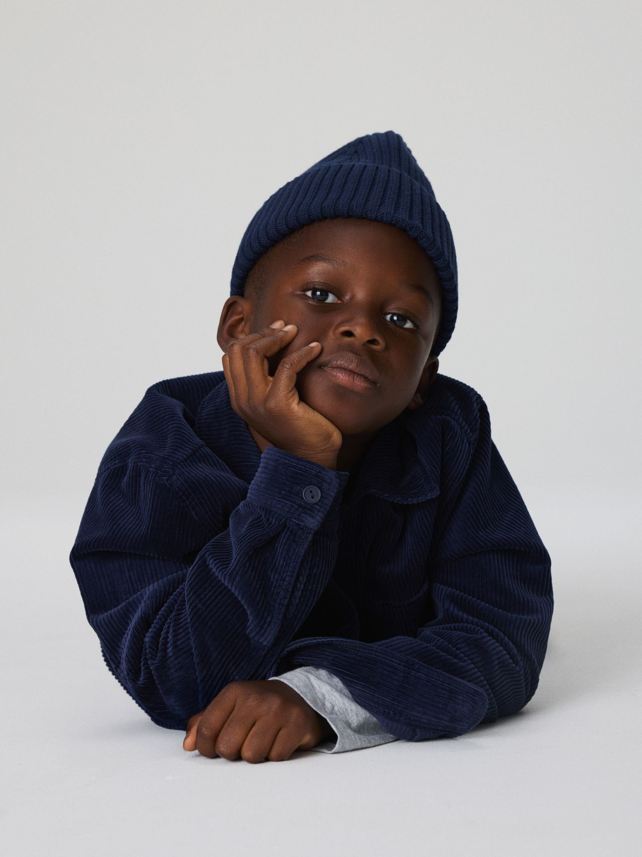 Child in a blue corduroy jacket and knit hat, resting chin on hand, gazing thoughtfully against a plain background.