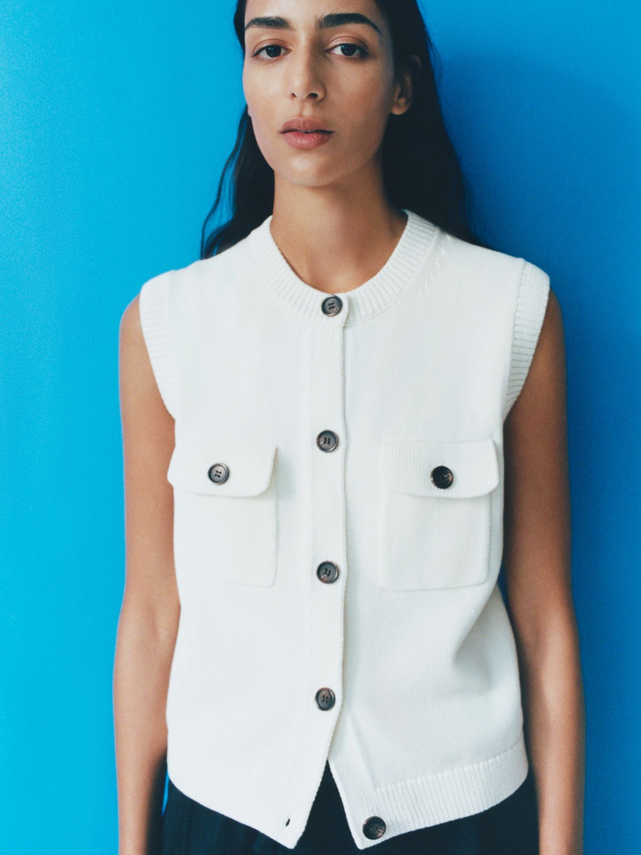 Person wearing a sleeveless white buttoned vest with pockets, standing against a bright blue background.