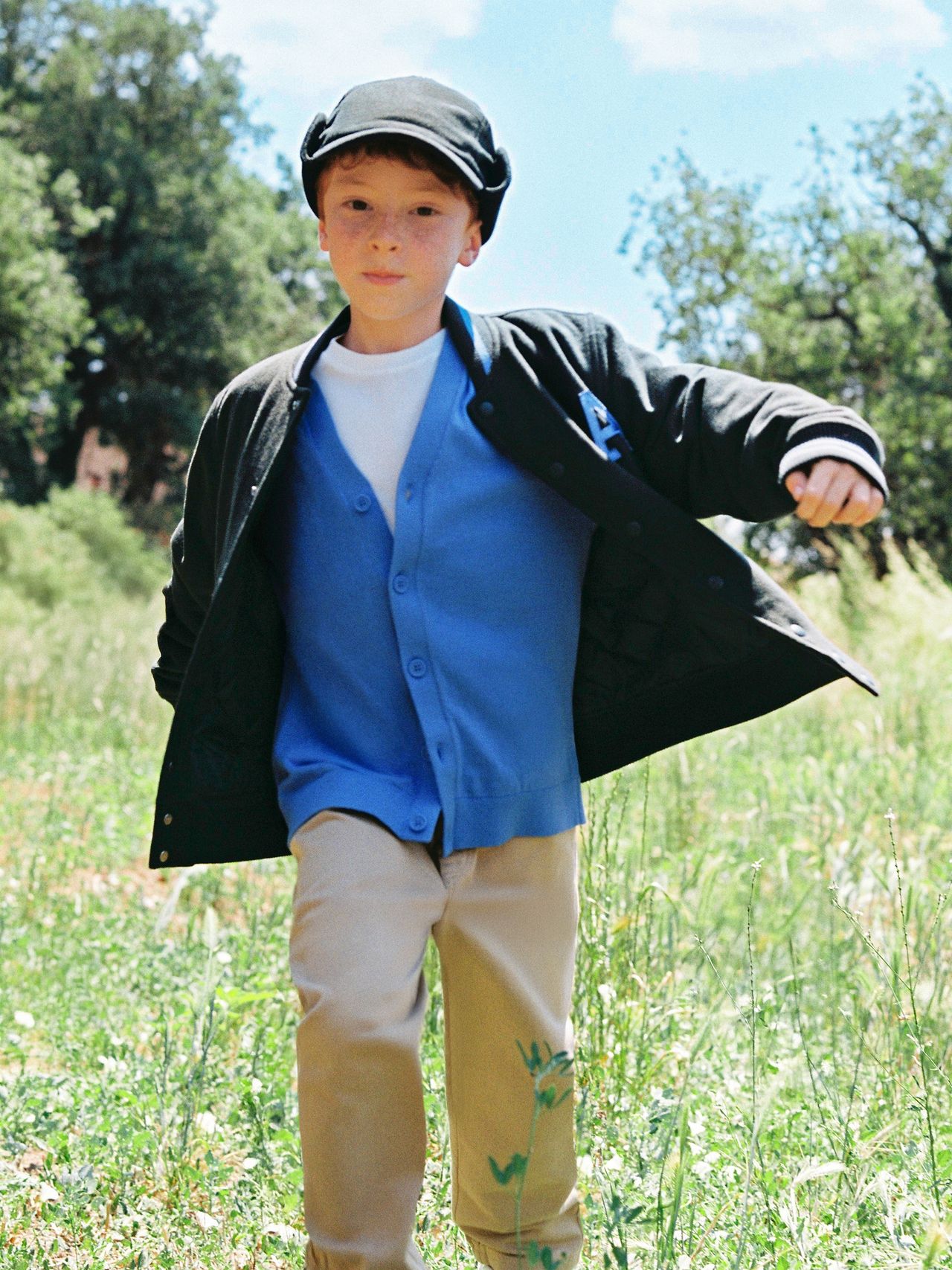 Young boy in a cap and blue cardigan runs through a sunny meadow, surrounded by trees and a clear sky.