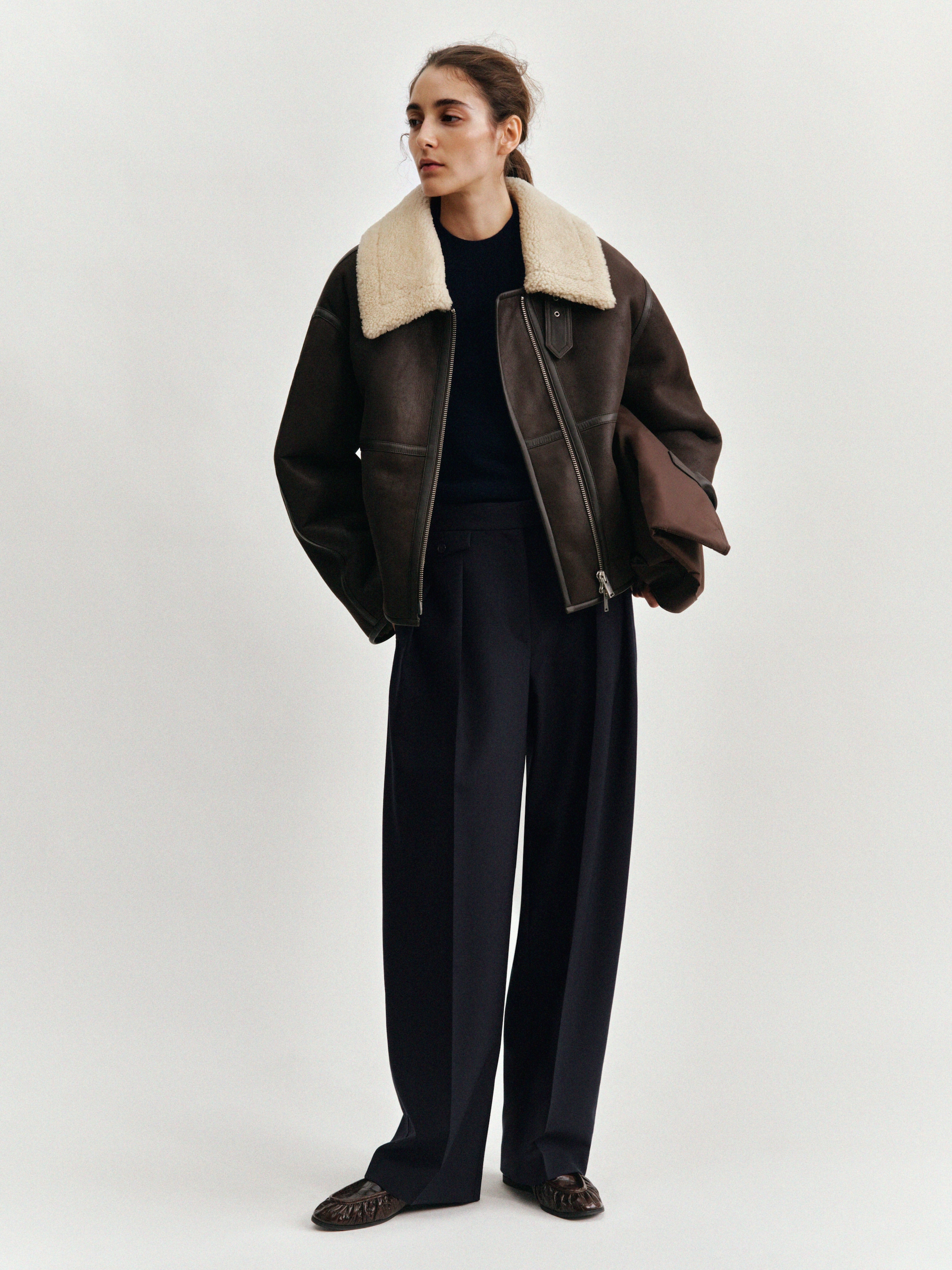 A person wearing a brown shearling jacket, dark pants, and brown shoes stands against a plain white background, looking to the side.