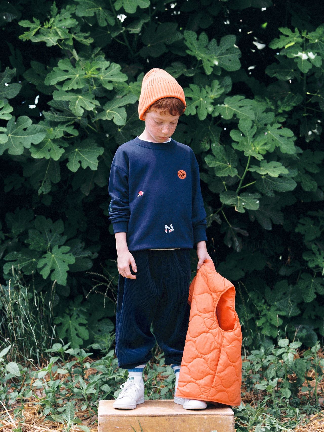 Young boy in a navy outfit and orange beanie stands on a box, holding an orange quilted vest, with lush green foliage in the background.