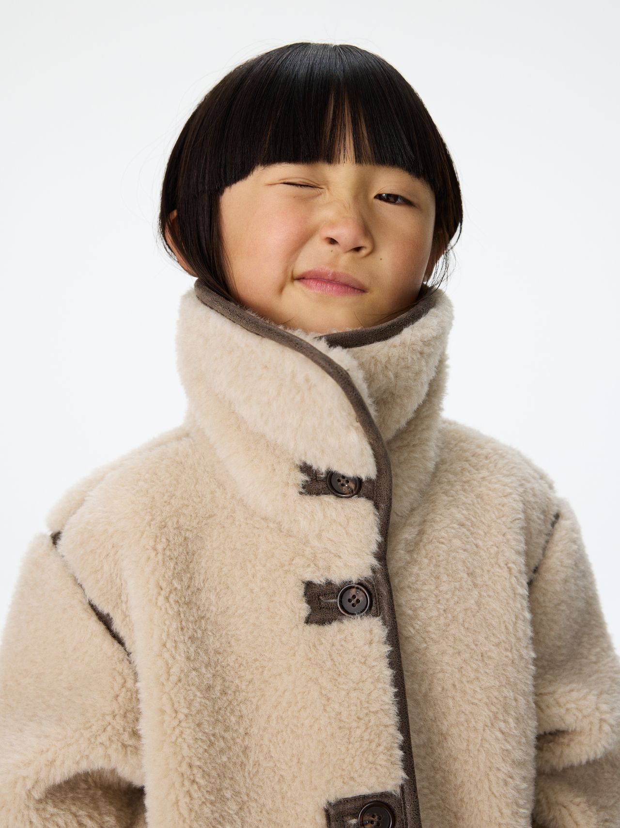 Child in a high-collared, beige fleece coat winks with a playful expression against a plain white background.