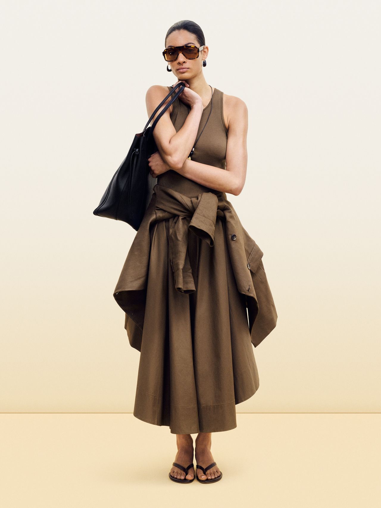 Woman in brown dress and sunglasses, holding a large black bag, with a jacket tied around her waist, standing against a plain background.