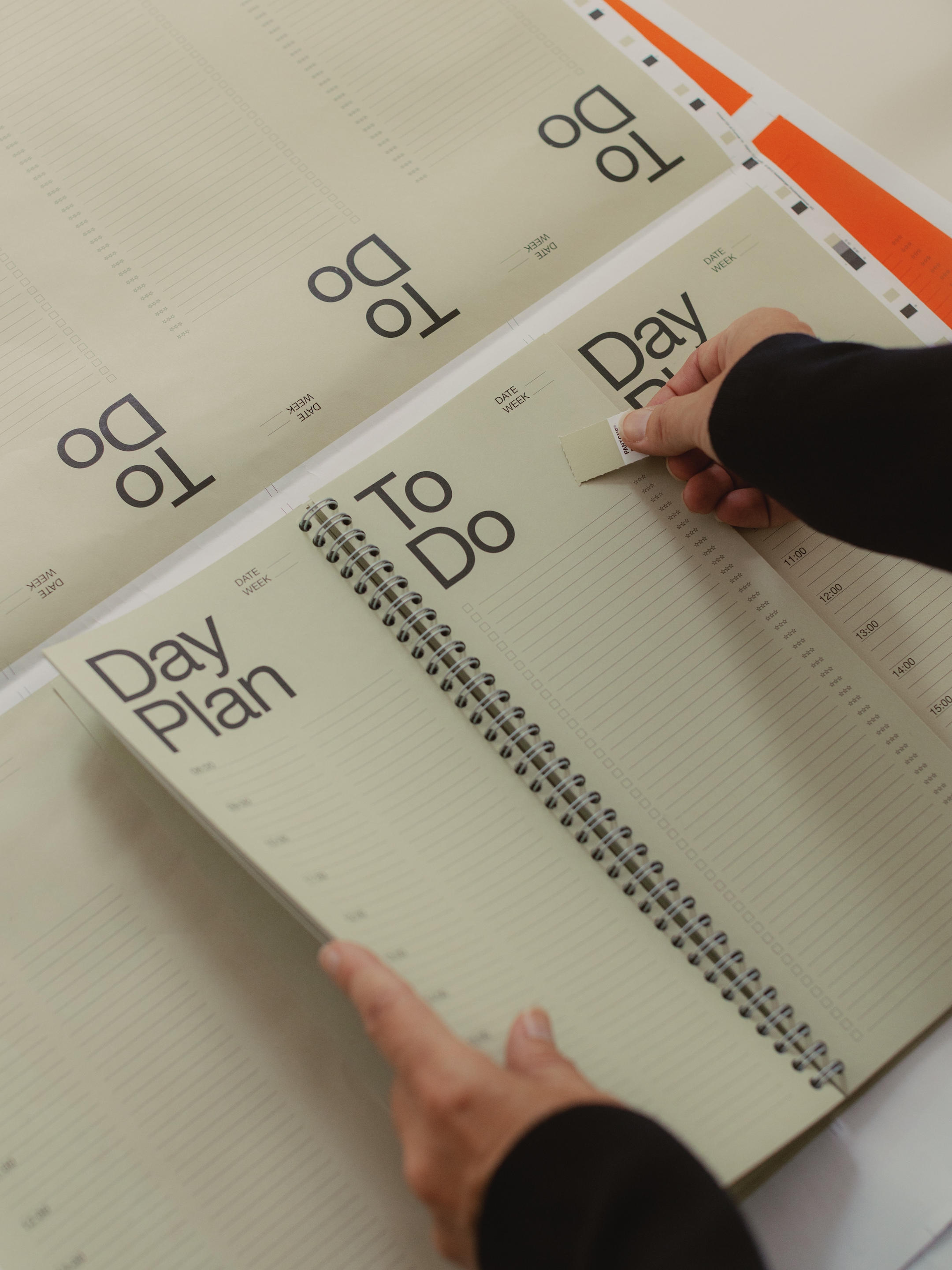 Person flipping through a spiral-bound planner with "Day Plan" and "To Do" headings on lined pages, checking off a task with a sticker.