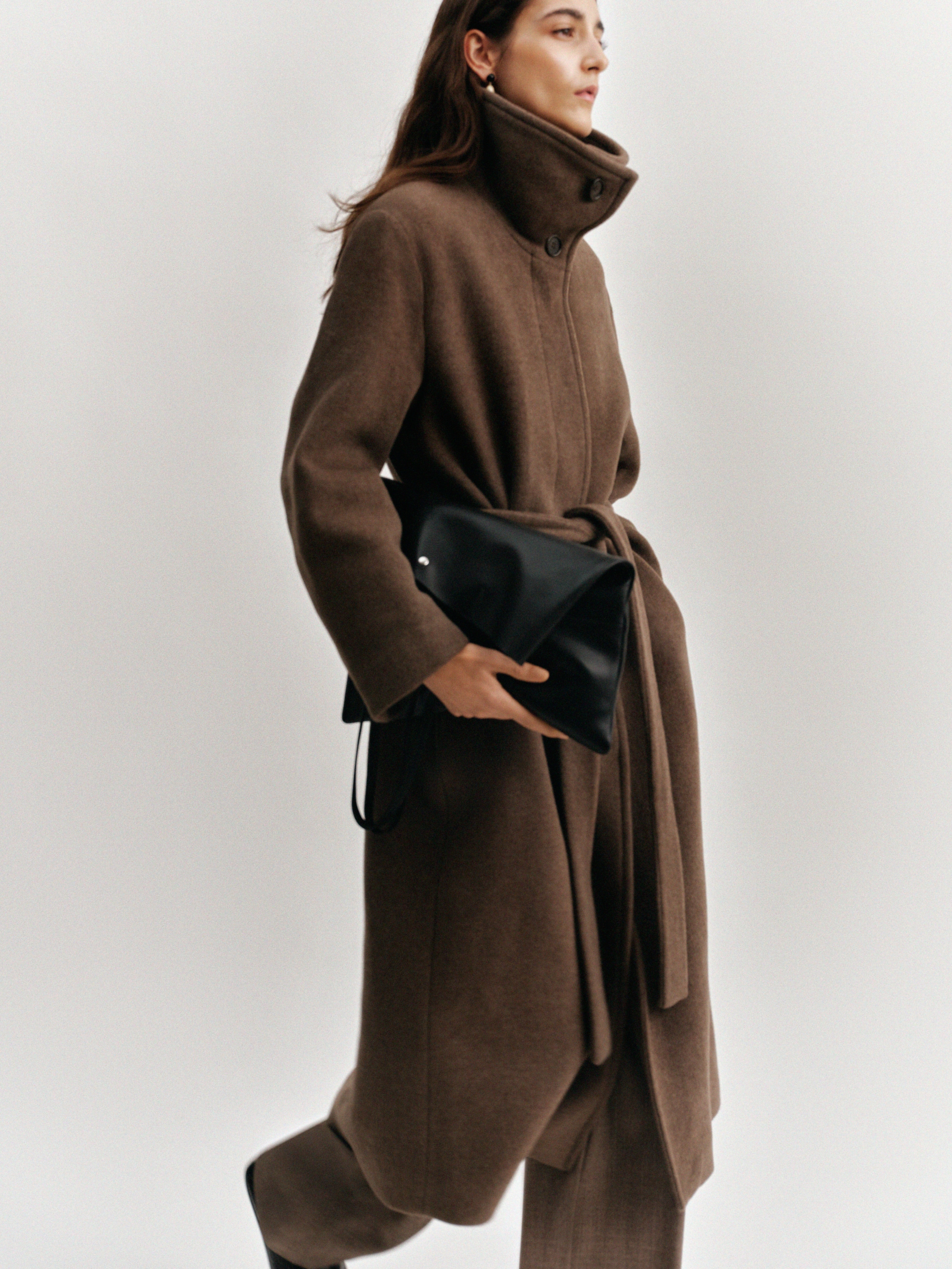 A person in a long brown coat with a high collar, holding a black handbag, walking against a plain background.