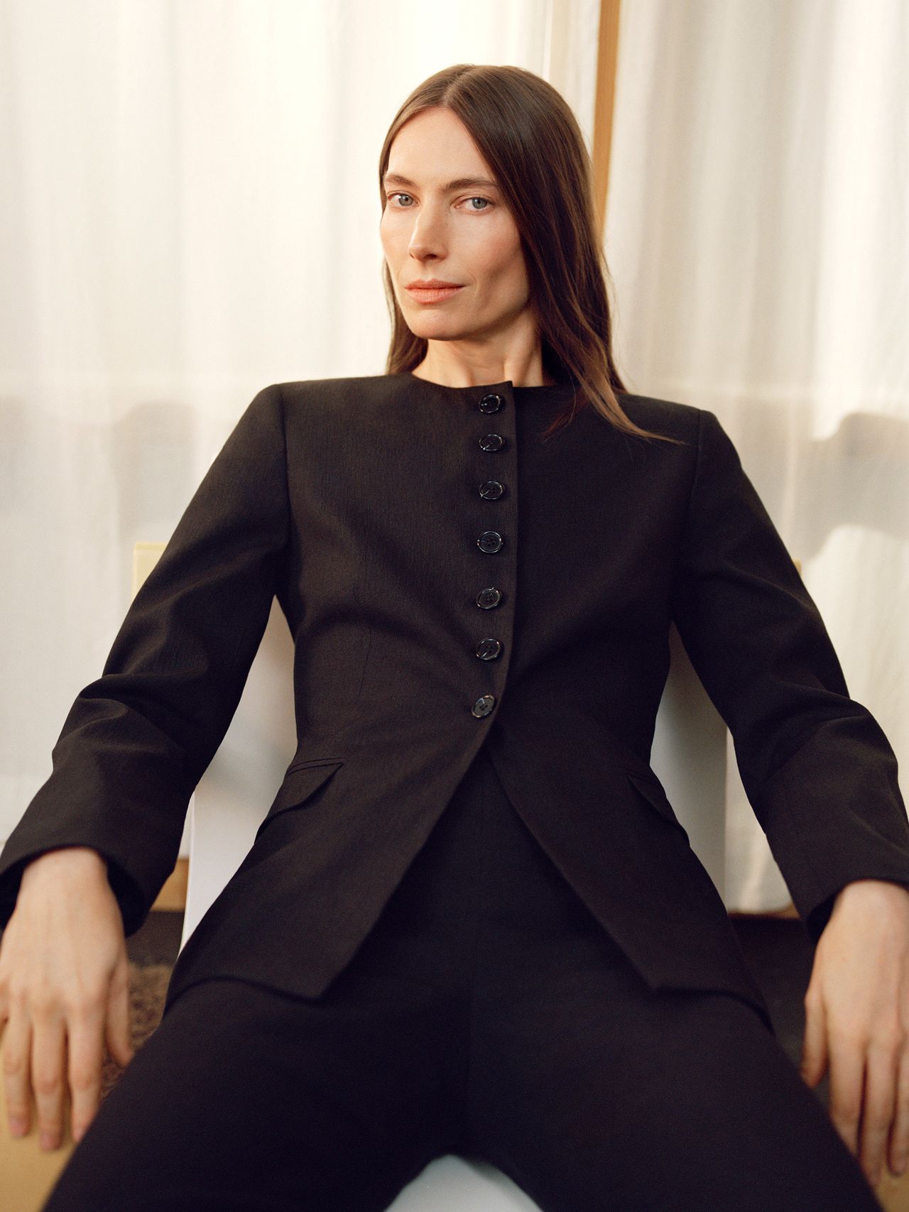 Person seated, wearing a tailored black suit, with long brown hair, in a relaxed pose against a light-colored curtain backdrop.