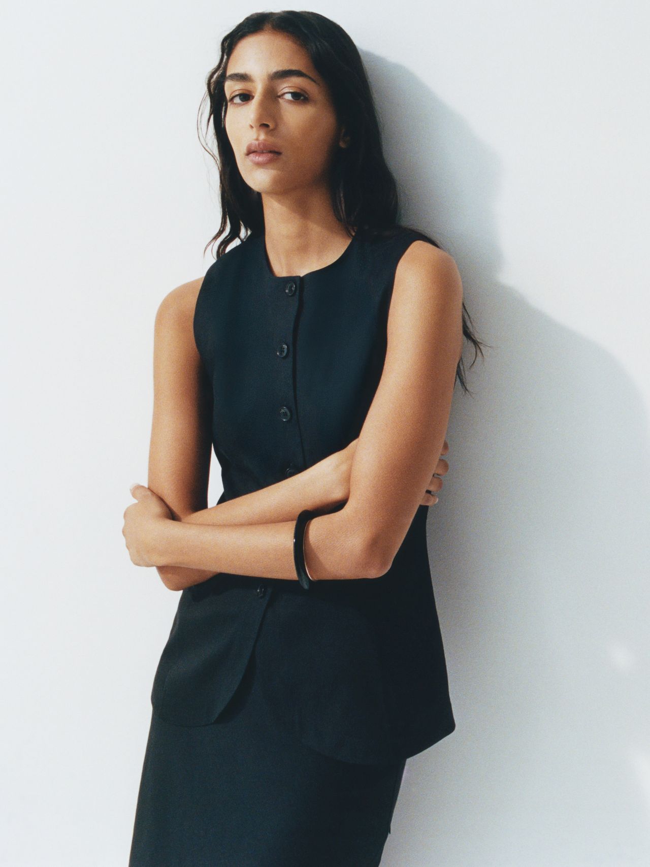 Person with long hair wearing a sleeveless black outfit, standing against a plain white background, arms crossed, with a neutral expression.