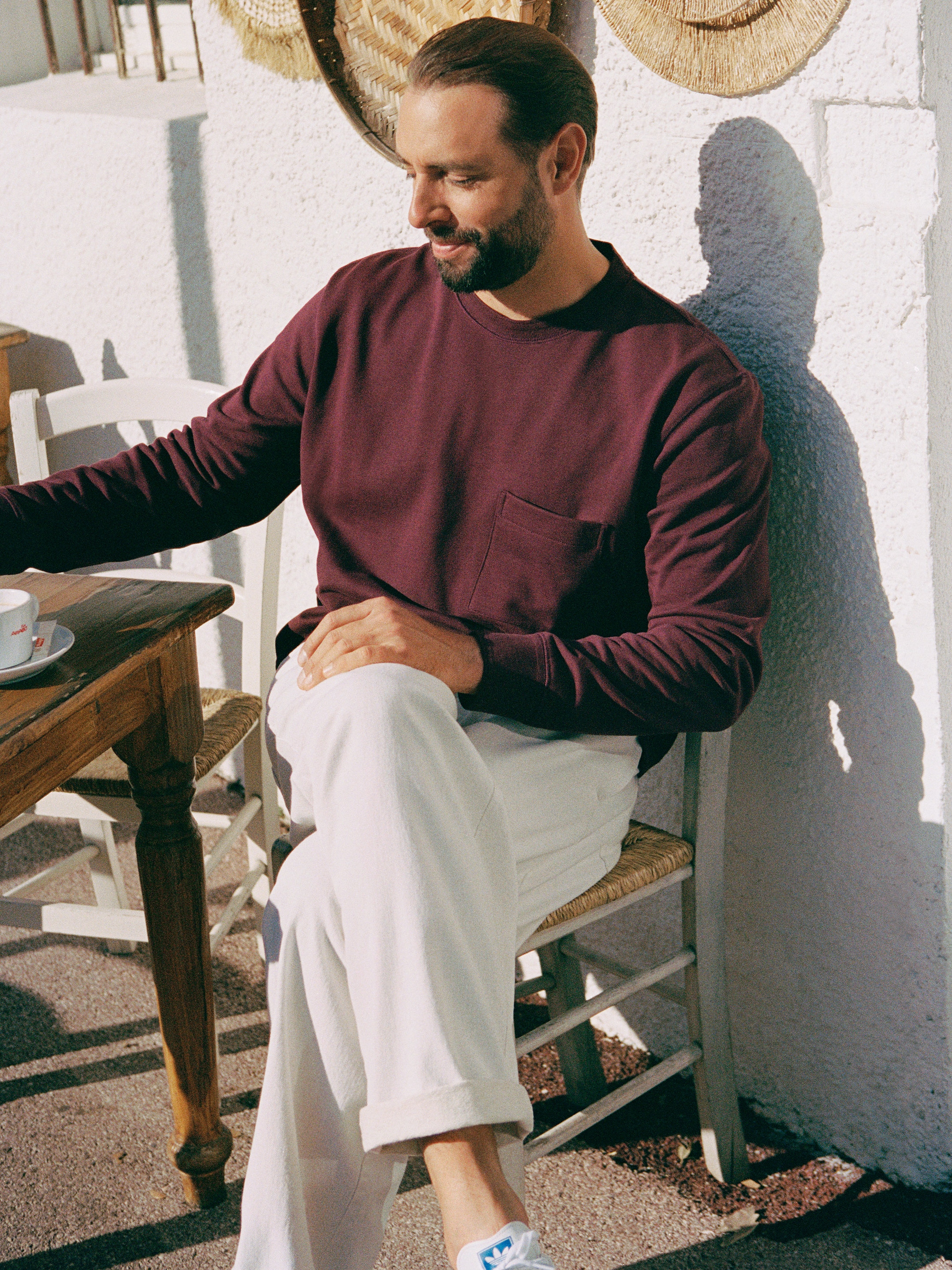 Man sitting outside at a wooden table, wearing a maroon sweater and white pants, holding a cup. Sunlight casts shadows on the wall.