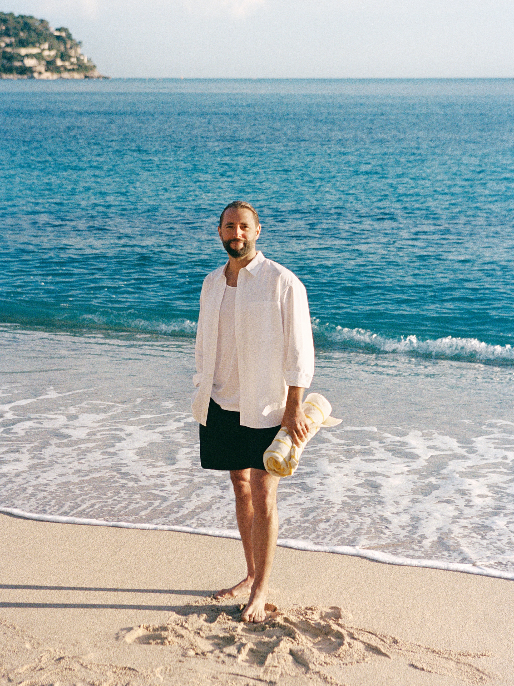 A man in a white shirt and black shorts stands barefoot on a sandy beach, holding a rolled-up towel, with the ocean in the background.